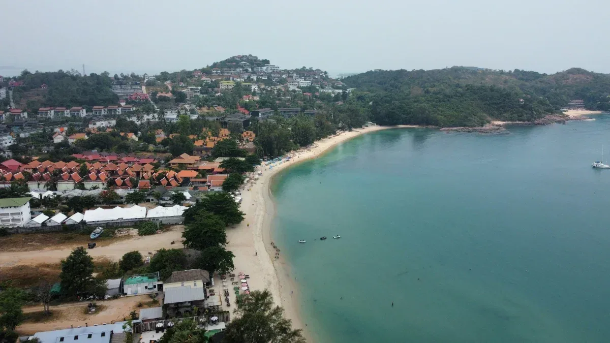 Aerial view of pristine curved bay in Koh Samui with turquoise waters, golden sandy beach, and resort buildings nestled in lush tropical forest