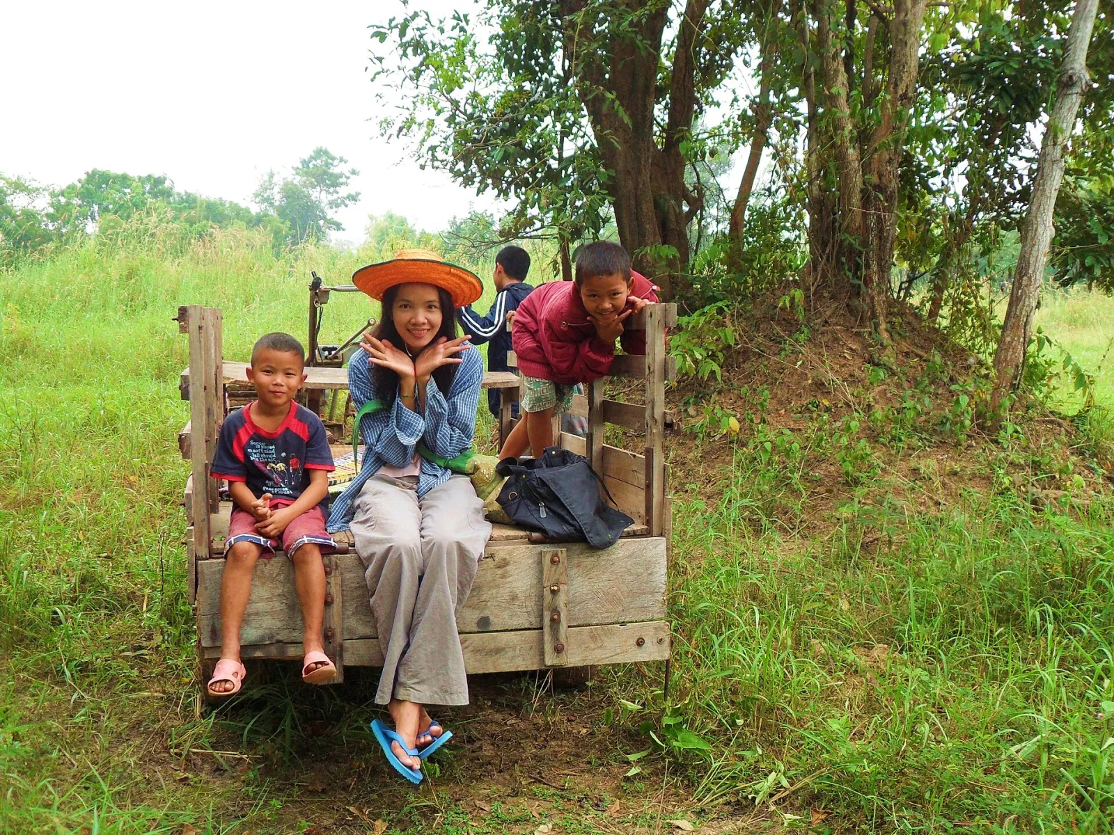 A woman in a straw hat and two children sit on a wooden cart in a grassy rural area, with other children playing in the background.