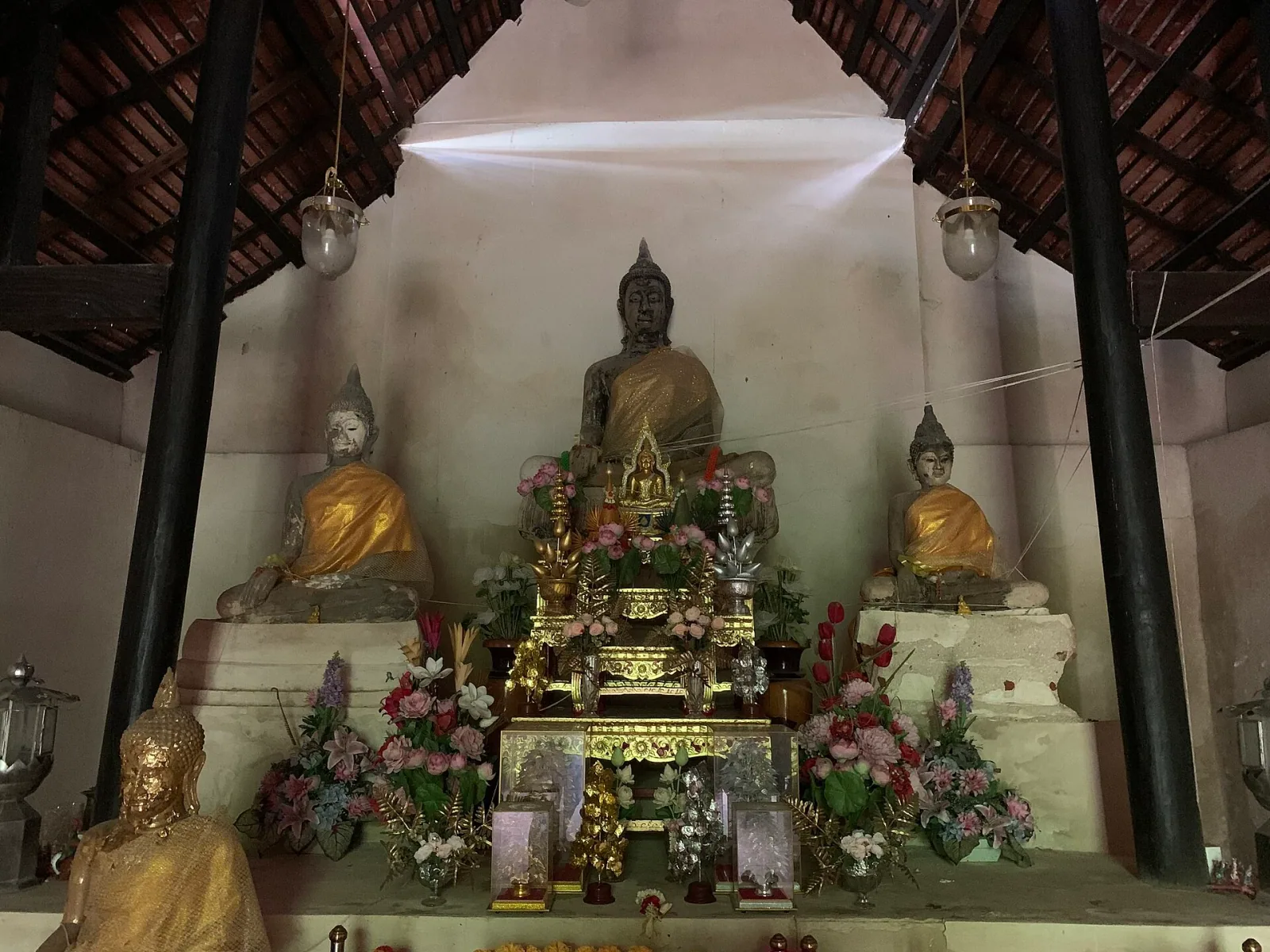 Interior of Wihan Luang Pho Kham at Wat Ban Pa in Ang Thong, featuring a large central Buddha statue flanked by two smaller ones, all adorned with golden robes and offerings.