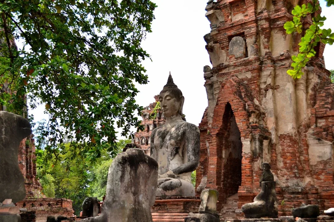A large, weathered stone Buddha statue sits in front of crumbling red brick temple ruins at Wat Phra Si Sanphet in Ayutthaya Historical Park, Thailand.