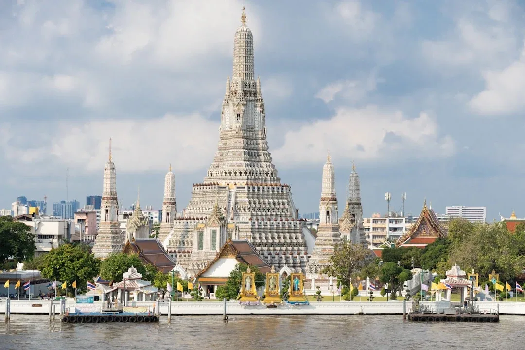 Panoramic view of Wat Arun, the Temple of Dawn, with its central prang and surrounding spires, seen from across the Chao Phraya River under a partly cloudy sky in Bangkok, Thailand.