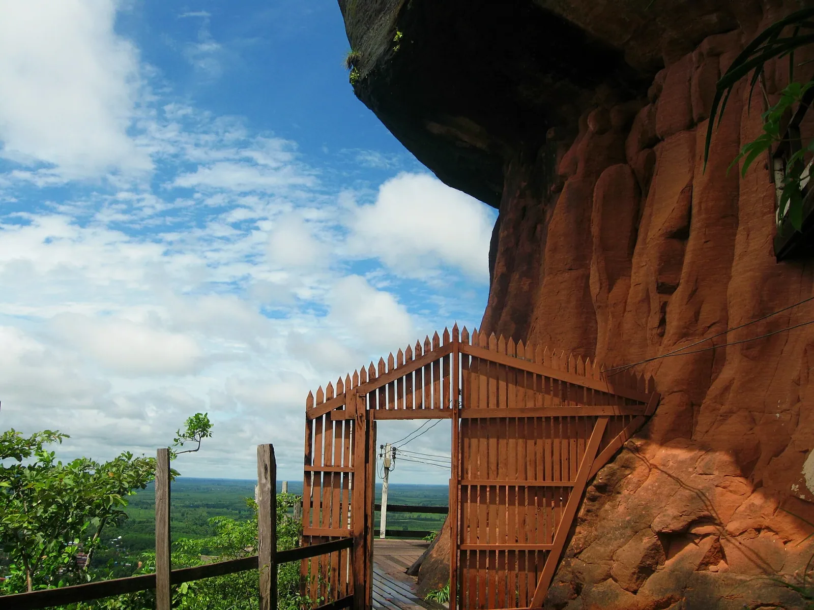 A wooden gate and walkway are integrated into a towering reddish-brown cliff, overlooking a vast green forest and distant horizon under a blue sky with white clouds at Phu Thok.