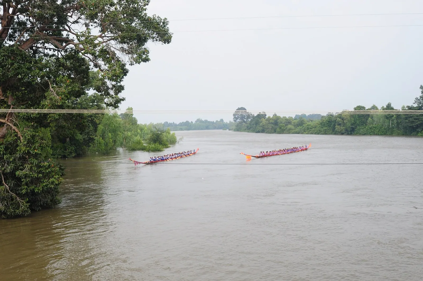 Two long, colorful boats filled with paddlers race on the Mun River, surrounded by green trees, during a festival in Buriram, Thailand.