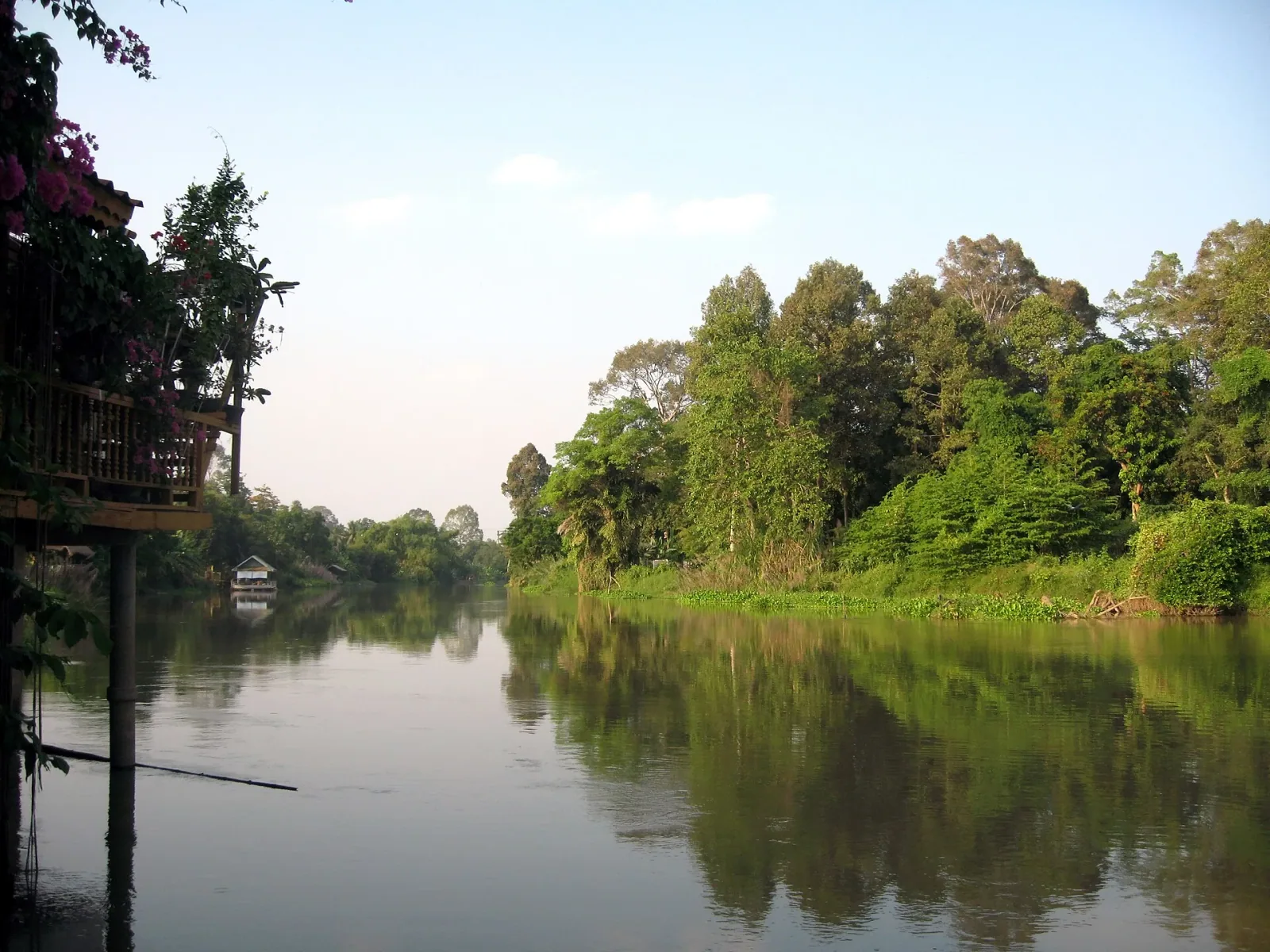 A tranquil river scene with dense green trees lining the far bank and a stilt house partially visible on the left, all reflecting in the calm water.