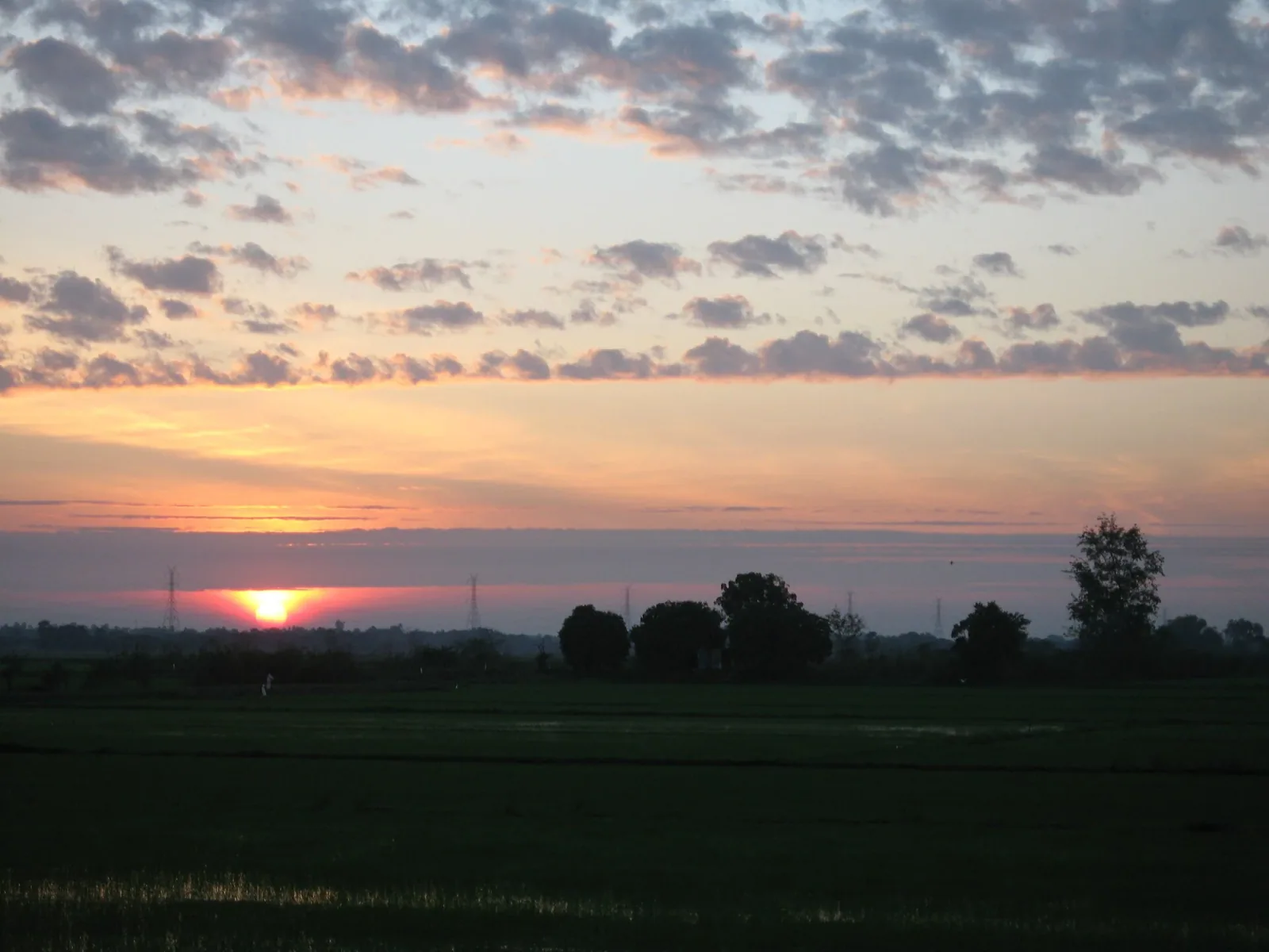 A vibrant sunrise or sunset with a bright orange sun low on the horizon, casting a glow over a flat rural landscape with silhouetted trees and dark fields under a cloudy sky.