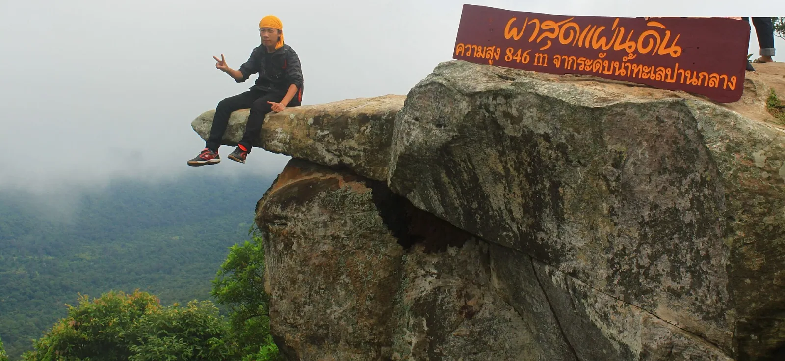 A person sits on a rock formation at Pha Sut Phaen Din viewpoint in Chaiyaphum, overlooking a misty, forested valley, with a sign indicating the location and altitude.