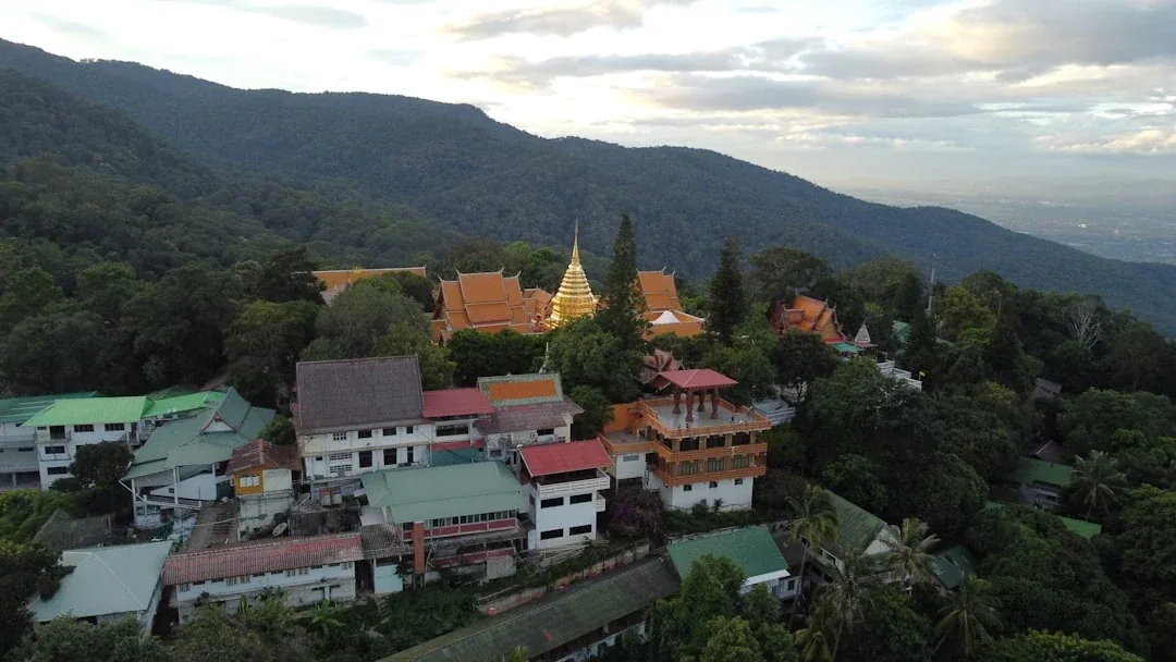 Aerial view of Wat Phra That Doi Suthep temple complex with its prominent golden chedi, surrounded by green forest on a mountain, overlooking a hazy valley in Chiang Mai, Thailand.