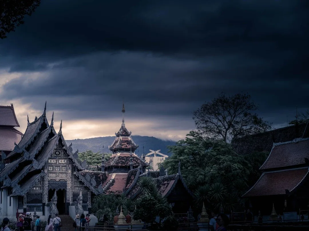 Traditional Thai temple with intricate dark roofs and spires, set against a dramatic, cloudy sky in Chiang Mai.