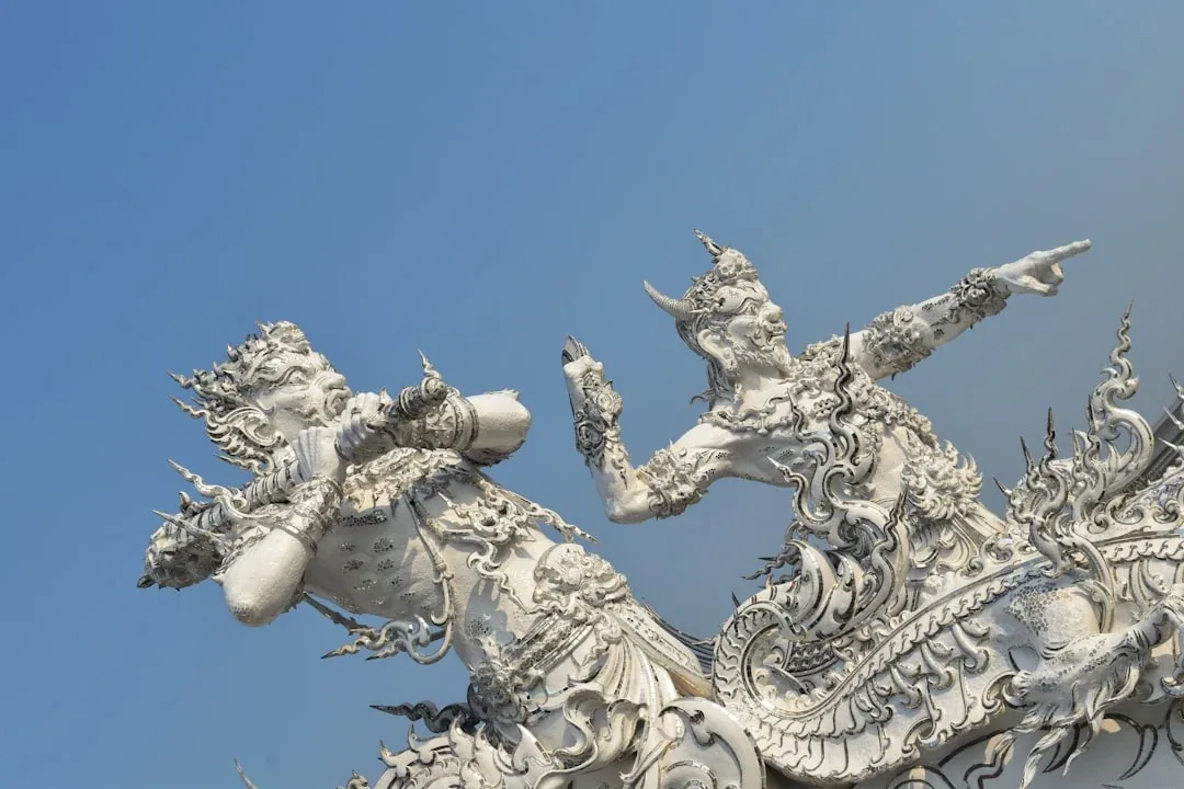 Close-up of two intricately carved white mythical guardian statues from Wat Rong Khun (White Temple) in Chiang Rai, Thailand, against a blue sky.