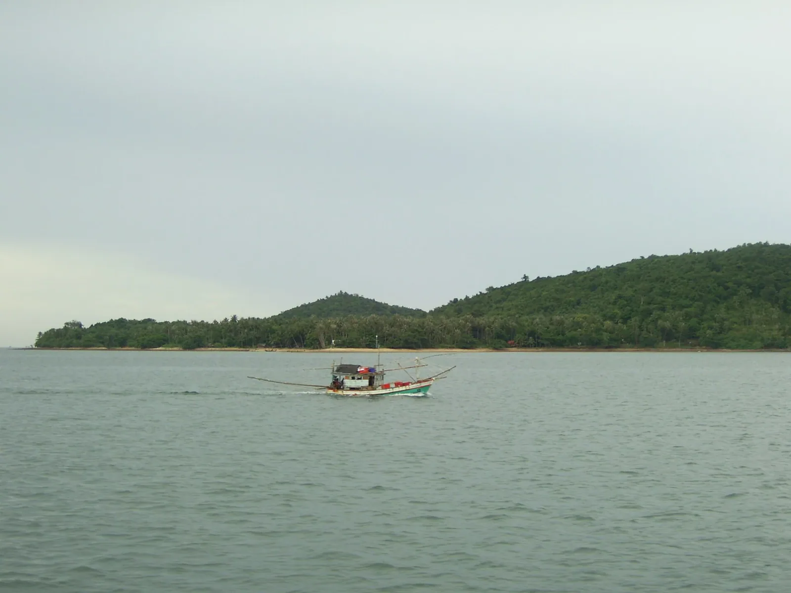 A small, colorful Thai fishing boat moves across calm waters with a forested island coastline in the background under an overcast sky.