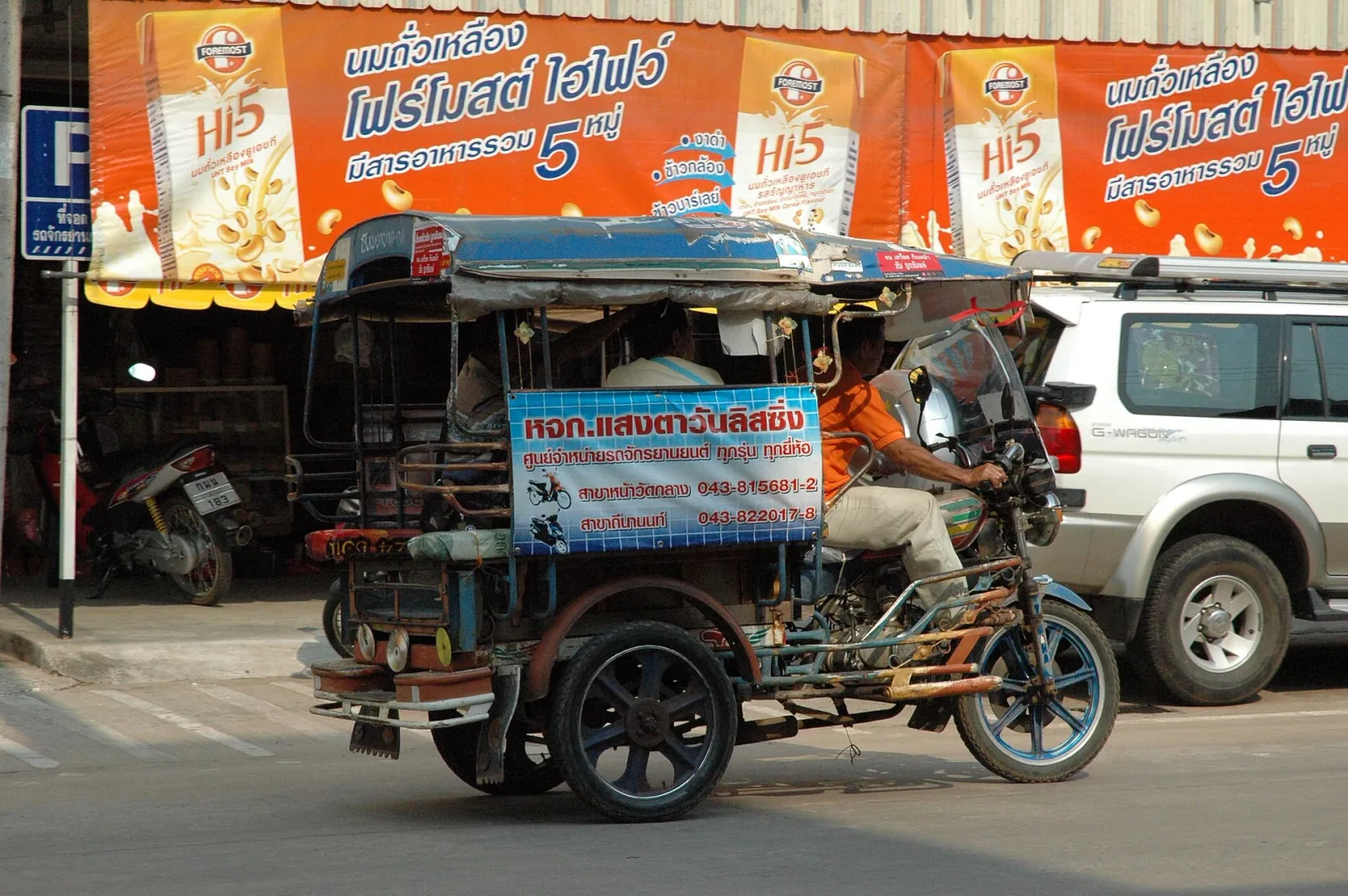A man drives a blue and red tuk-tuk on a street in Thailand, with a white SUV and storefront banners in the background.