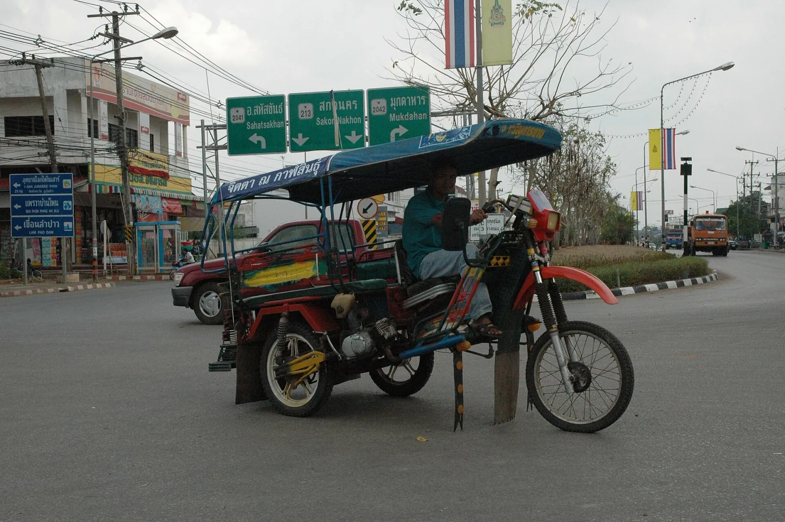 A man drives a red and blue dirt bike-converted tuk-tuk on a street in Kalasin, Thailand, with green road signs in the background pointing to local districts and other provinces.