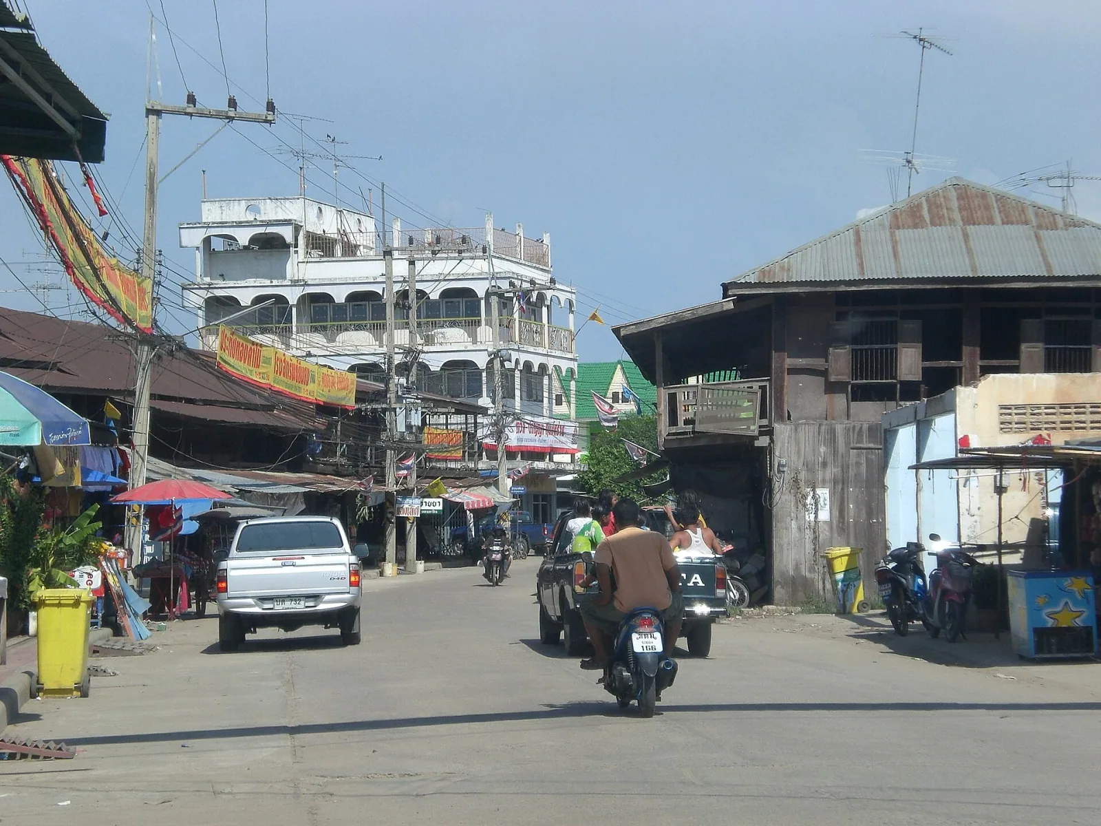 A busy street in a Thai town, with market stalls and shops lining the road, various motorbikes and a silver pickup truck driving, and a mix of old wooden and multi-story concrete buildings under a bright sky.