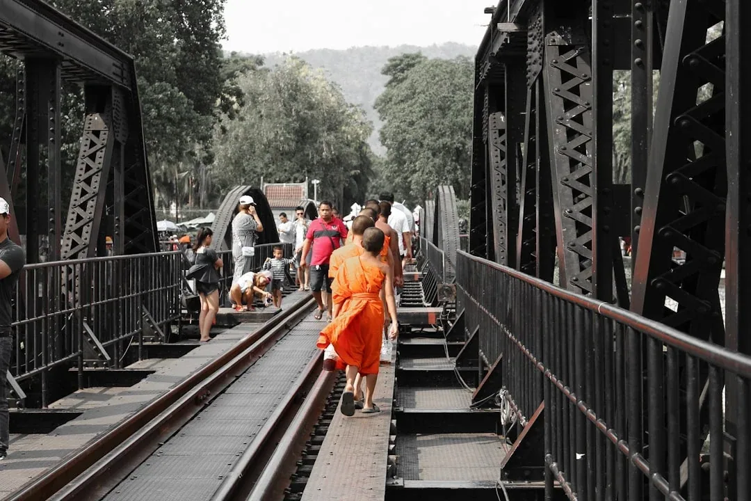 People, including Buddhist monks in orange robes, walk along the railway tracks of the black metal Bridge on the River Kwai in Kanchanaburi, Thailand.
