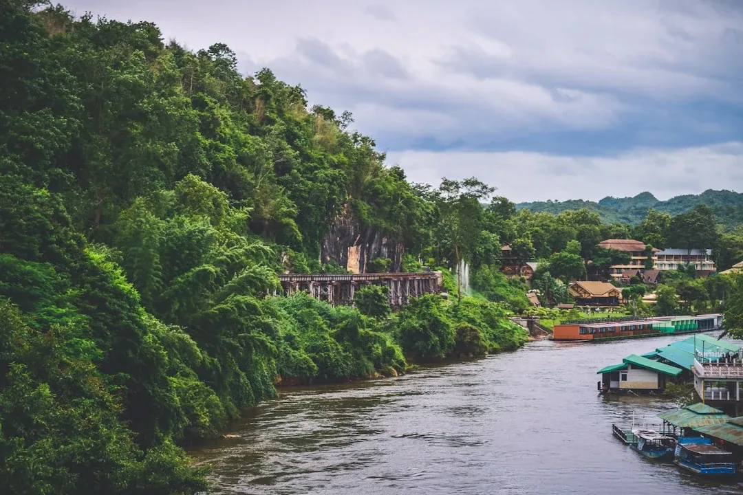 Scenic view of the Kwai River in Kanchanaburi, Thailand, with the historic Death Railway viaduct built into a lush green cliff and floating houses on the water.