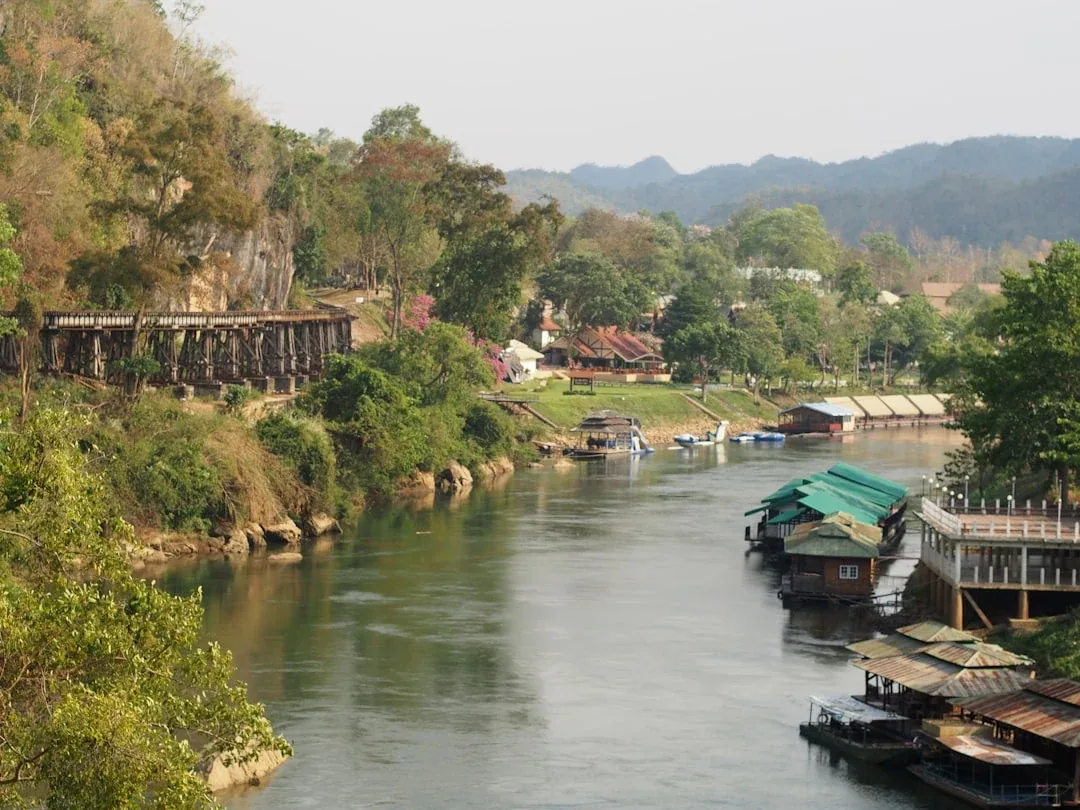 Panoramic view of the Kwai River in Kanchanaburi, Thailand, showing the historic wooden viaduct of the Death Railway on the left bank and floating houses on the right, surrounded by lush hills.