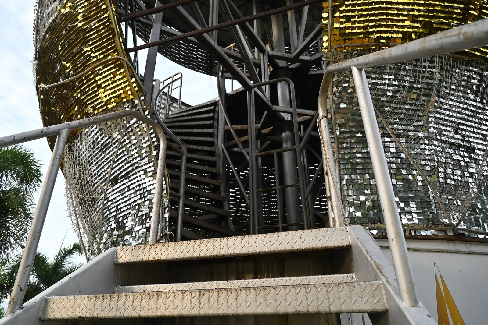 A close-up of a metal staircase with diamond plate treads leading up to the base of the Phra Mahathat Kaen Nakhon stupa, which is covered in reflective gold and silver panels, in Khon Kaen.