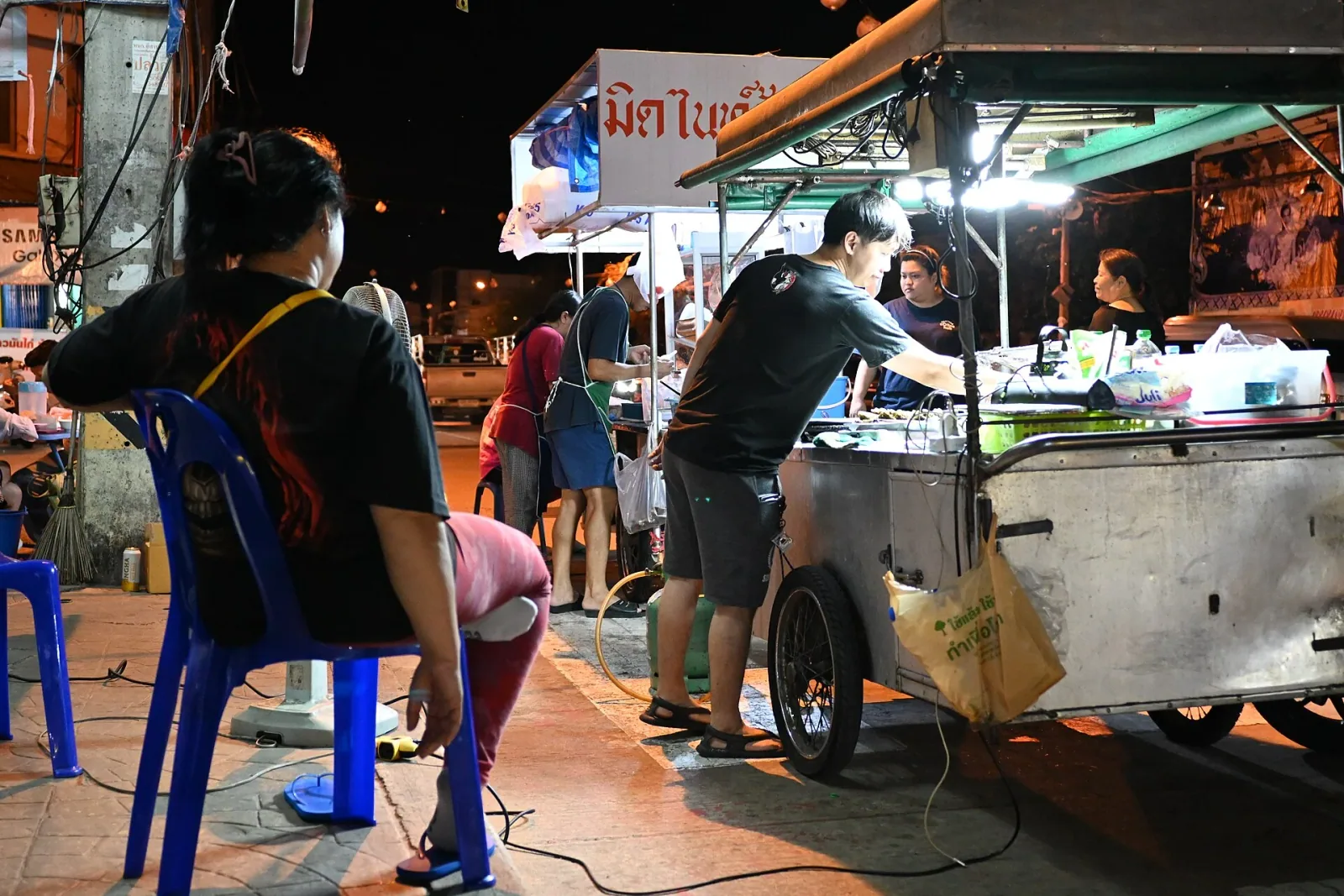 A brightly lit street food cart at night in Thailand, with a vendor preparing food and customers gathered around, including a woman seated in the foreground.