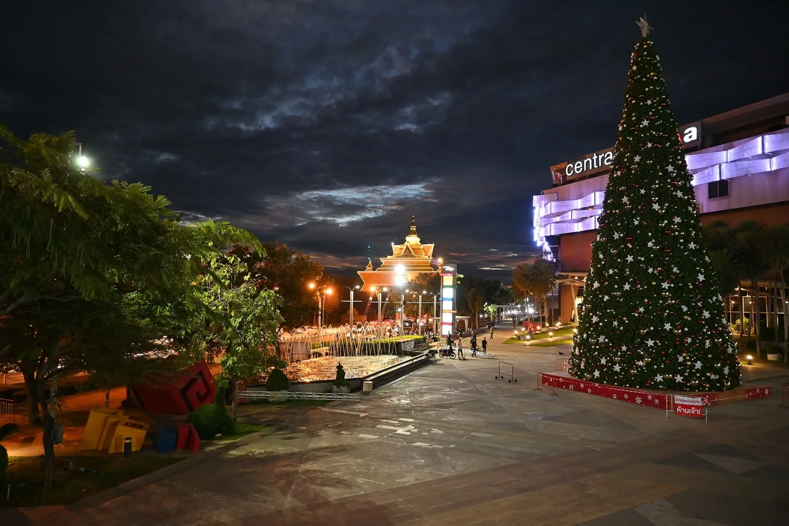 A nighttime view of CentralPlaza Khon Kaen's outdoor plaza, featuring a large, brightly lit Christmas tree, a modern shopping complex with purple lighting, and an ornate Thai-style pavilion in the background.