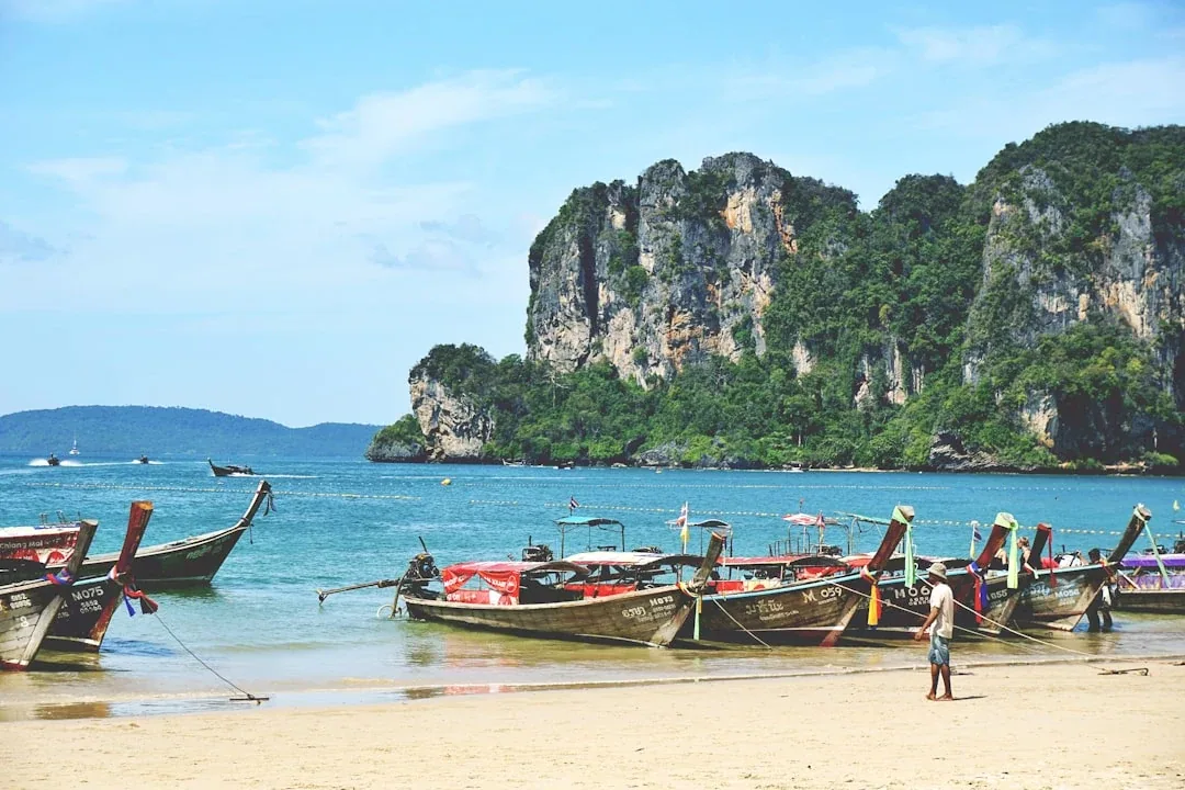 Longtail boats moored on the sandy shore of Railay Beach in Krabi, Thailand, with towering, jungle-covered limestone cliffs rising from the turquoise sea in the background.
