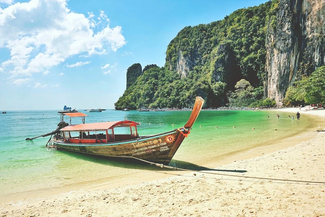 A traditional Thai longtail boat with a red canopy moored on a sandy beach with clear turquoise water, backed by towering limestone cliffs covered in green foliage under a blue sky.
