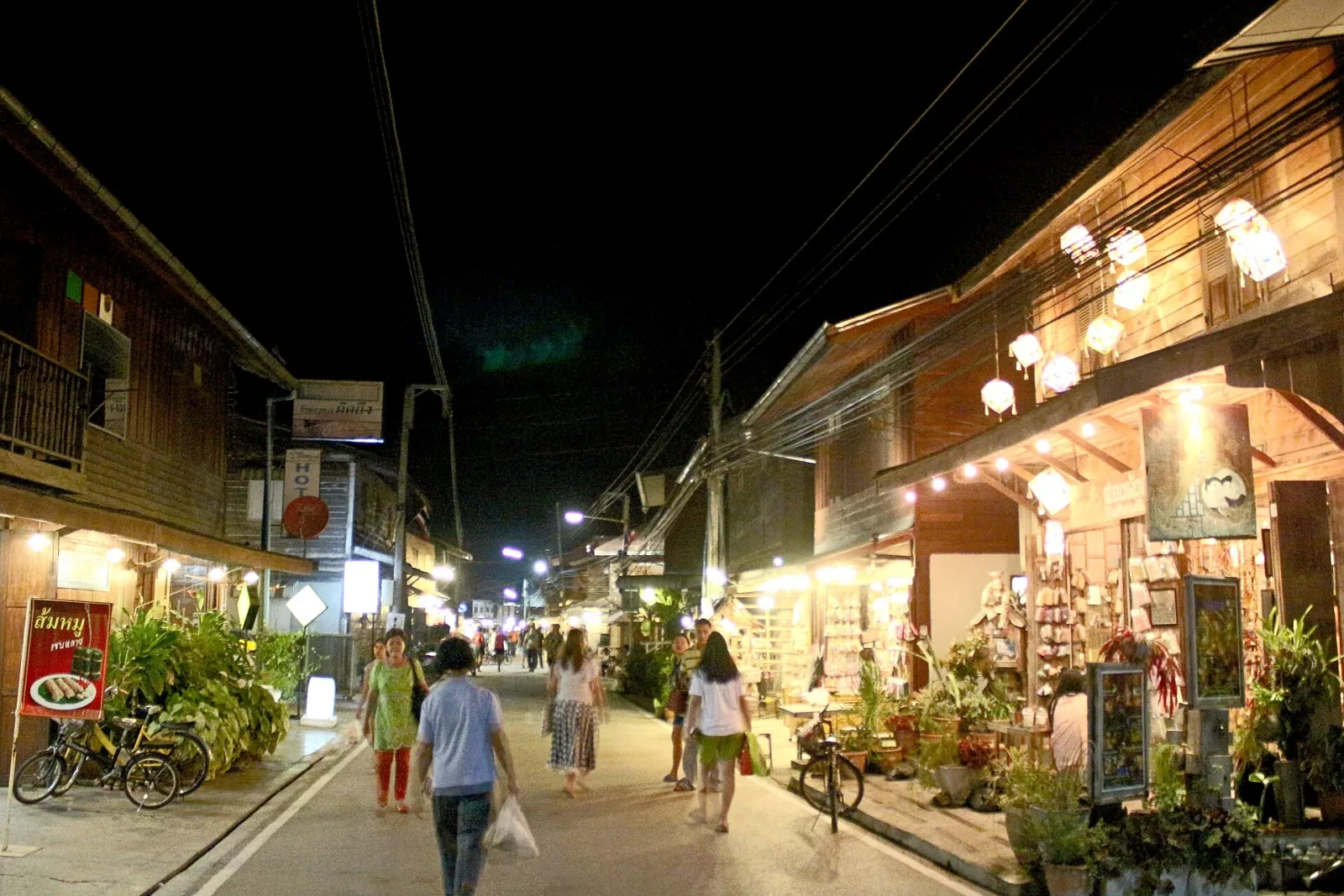 A bustling night market street in Chiang Khan, Loei, Thailand, with traditional wooden shophouses lit by warm lights, and people walking along the street.