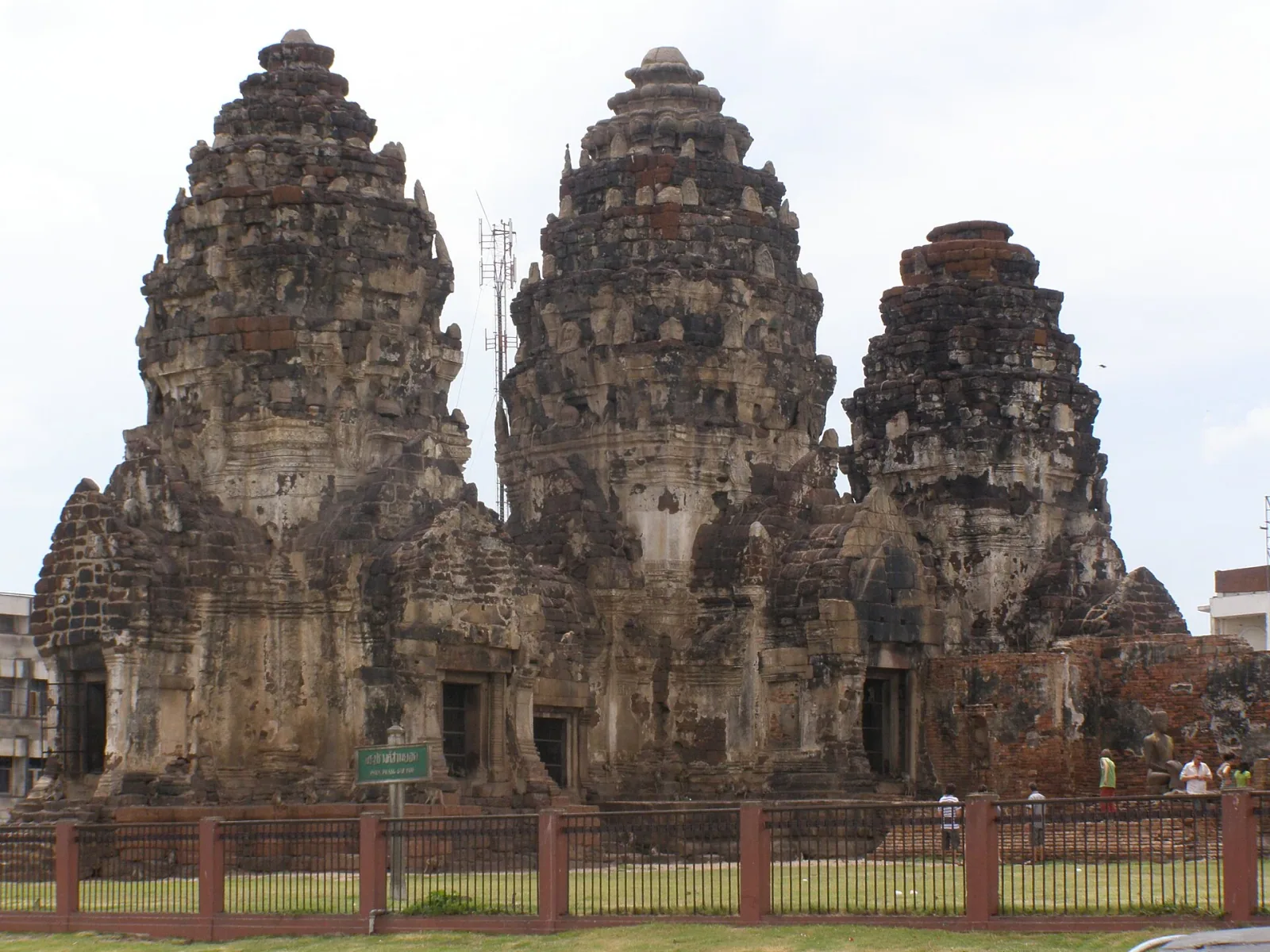 Phra Prang Sam Yot, an ancient Khmer-style temple in Lopburi, Thailand, featuring three weathered laterite towers under a bright sky, with a fence in the foreground.