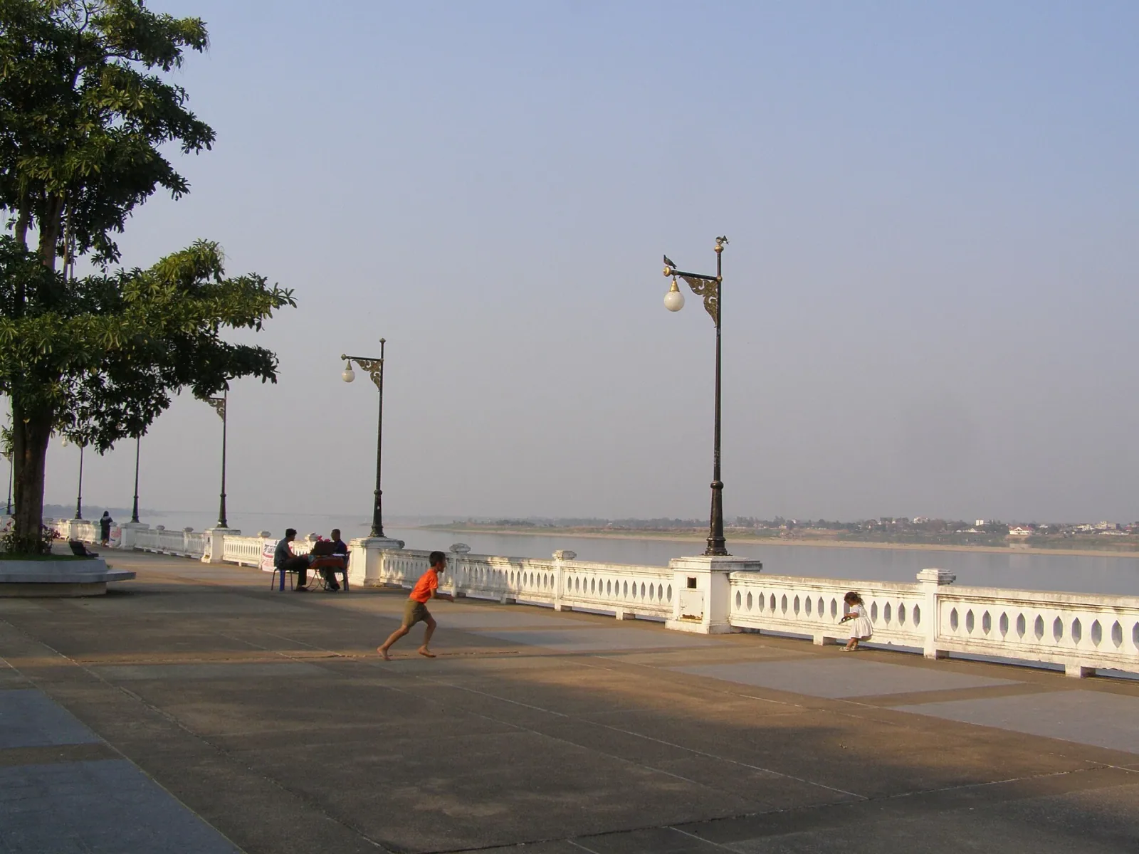 A child runs barefoot on a riverside promenade in Mukdahan, Thailand, with the Mekong River and distant Laos visible under a hazy sky.