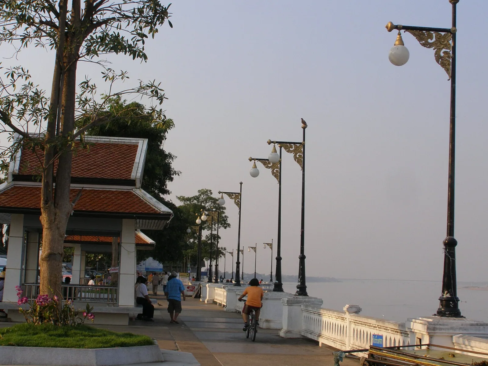 A riverfront promenade in Mukdahan with a traditional Thai pavilion, ornate lampposts, and people walking and cycling along the Mekong River.