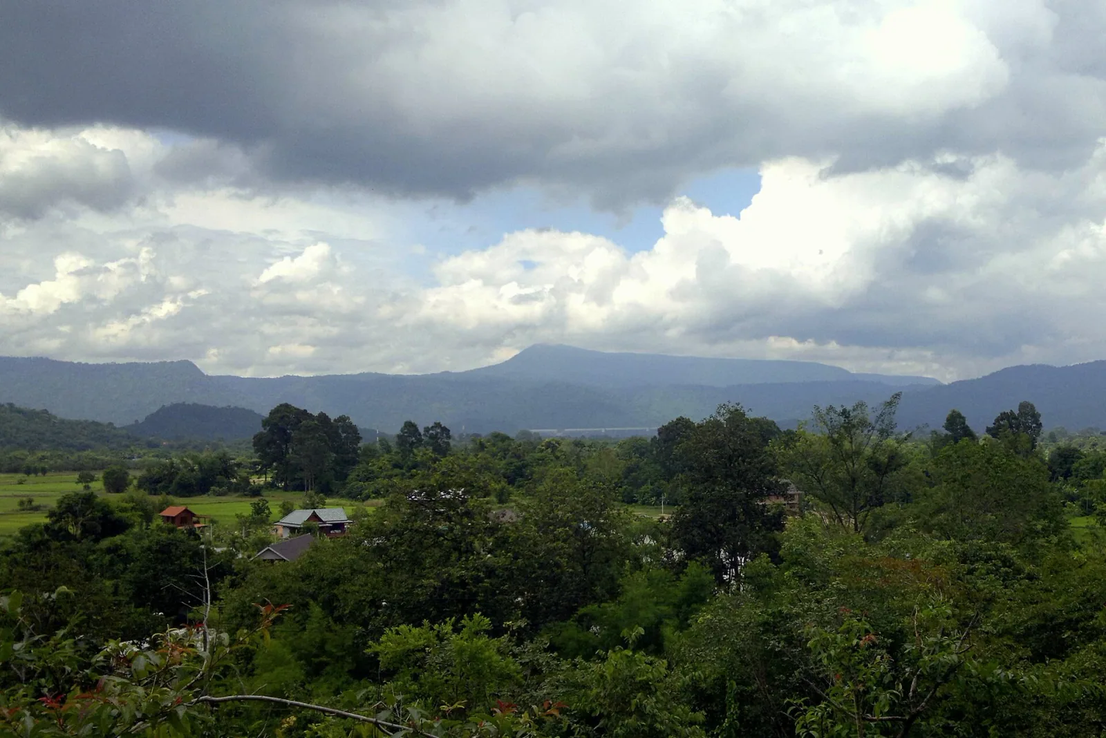Panoramic view of Khao Lon mountain range in Nakhon Nayok, with lush green trees, fields, and a few houses in the foreground under a cloudy sky.