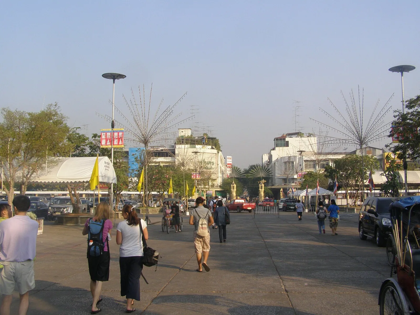 A wide street or plaza in Nakhon Pathom, Thailand, with pedestrians walking, parked cars, and buildings, leading towards the golden Phra Pathom Chedi in the background.
