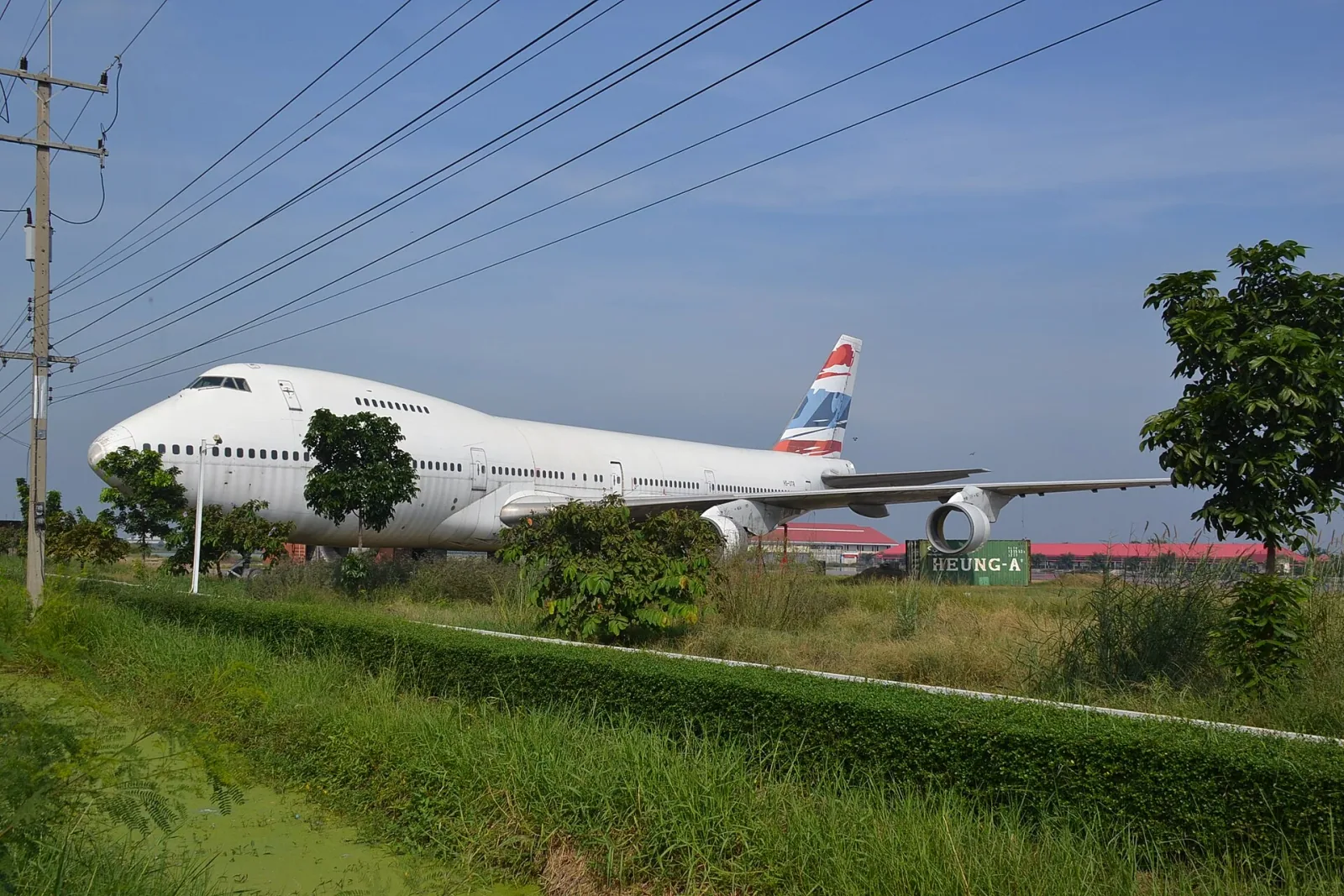 A decommissioned white Boeing 747 airplane with a red, white, and blue striped tail sits in a grassy field next to a road under power lines in Nakhon Pathom.