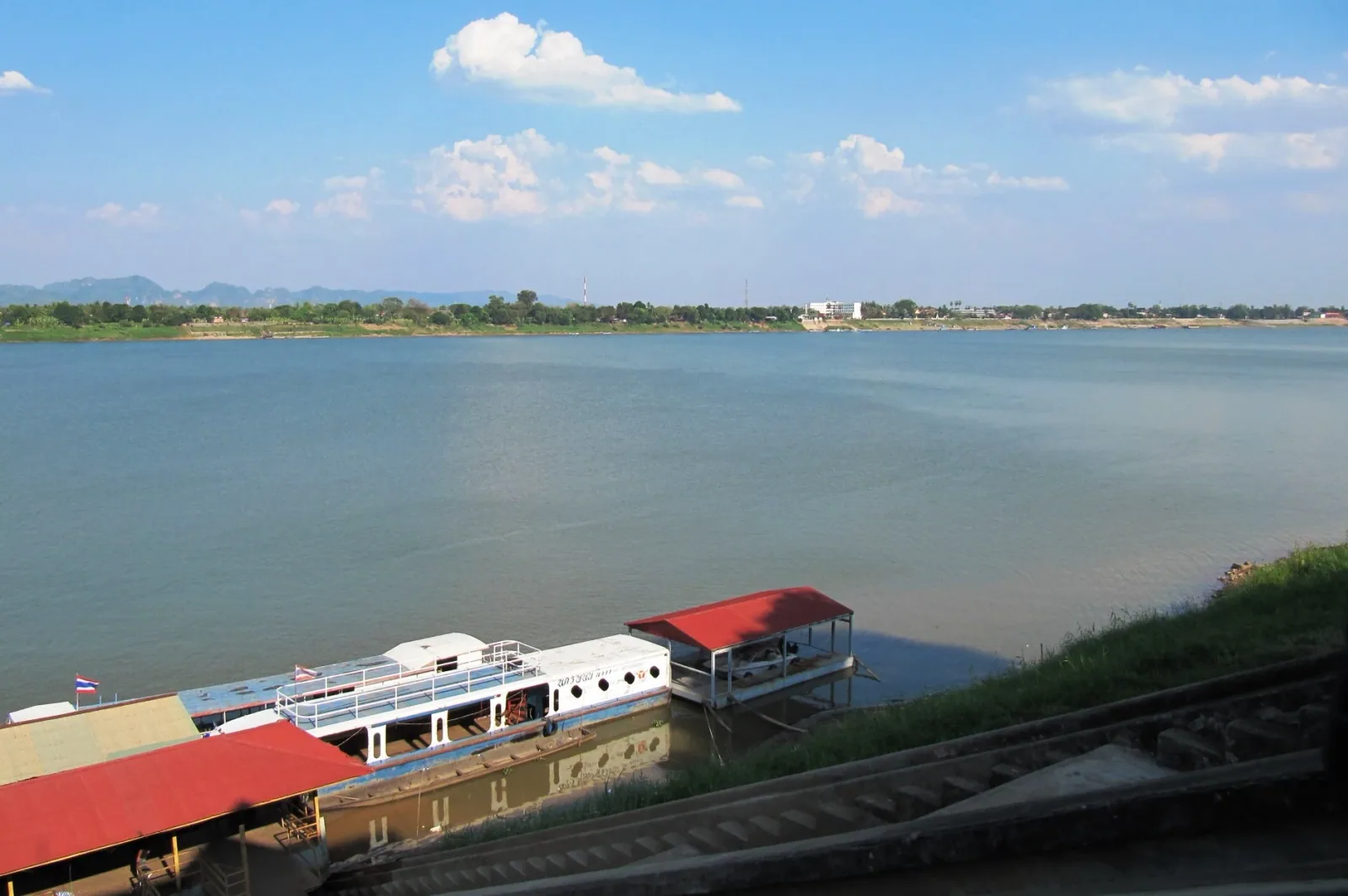 Panoramic view of the wide Mekong River from Nakhon Phanom, Thailand, with two docked boats in the foreground and the distant green shore of Thakhek, Laos, under a blue sky.