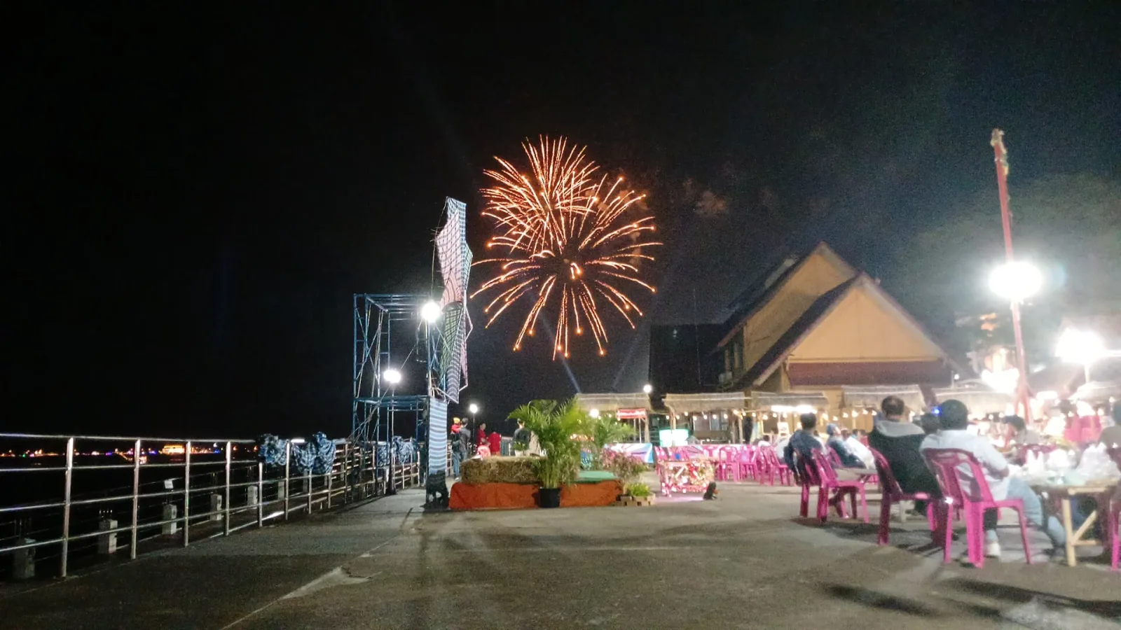 Fireworks illuminate the night sky over a bustling waterfront promenade in Nakhon Phanom, with people enjoying the Chinese New Year festival at tables.