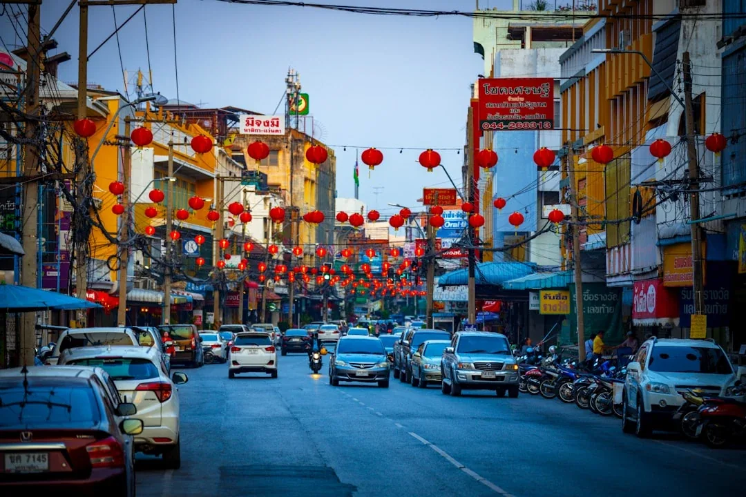 A bustling street in Nakhon Ratchasima city, Thailand, adorned with numerous red Chinese lanterns during Chinese New Year, with cars and motorcycles on the road and buildings lining both sides.