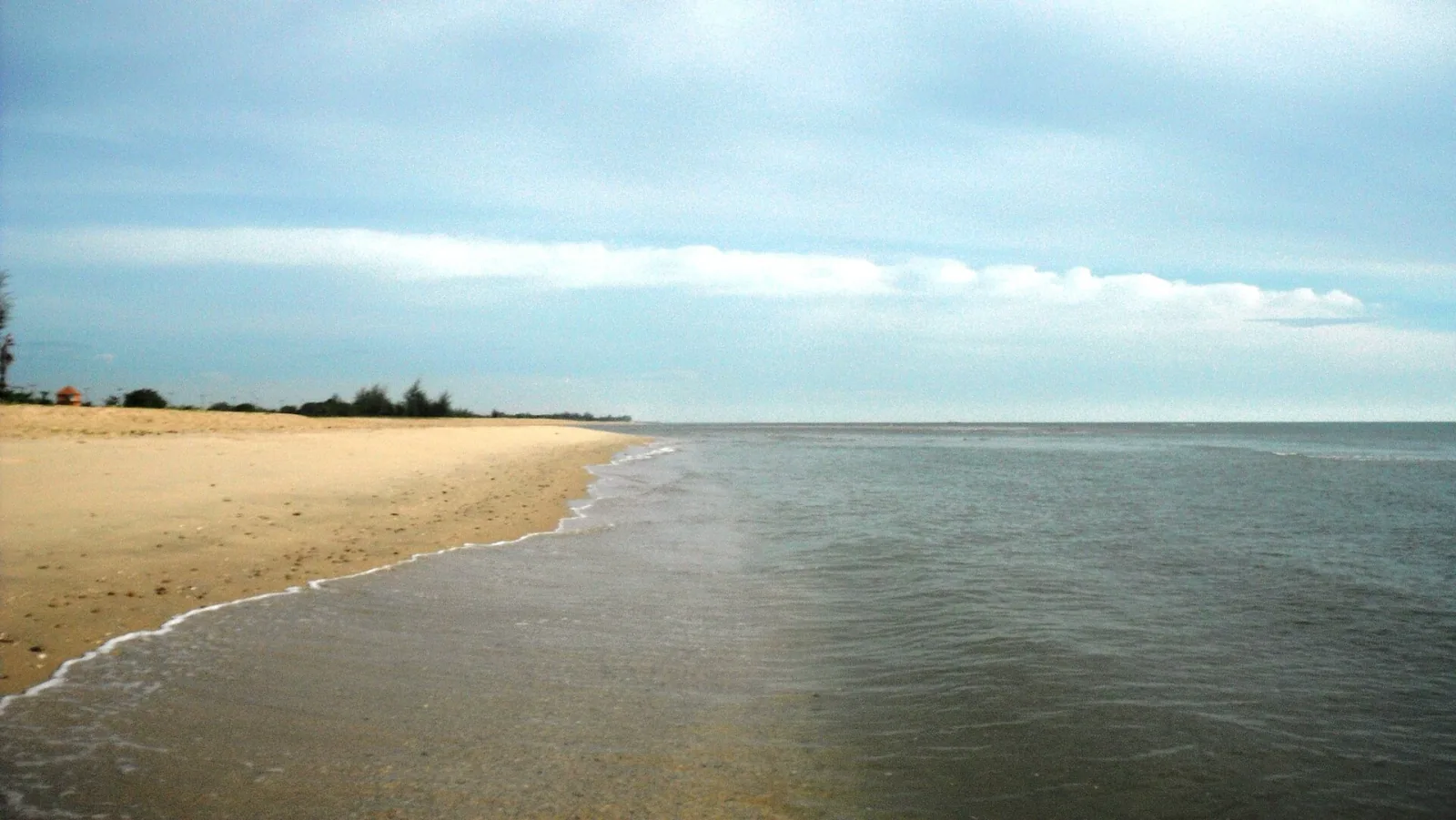 A wide sandy beach with small waves breaking on the shore, stretching towards a distant tree line under a light blue sky with scattered clouds.