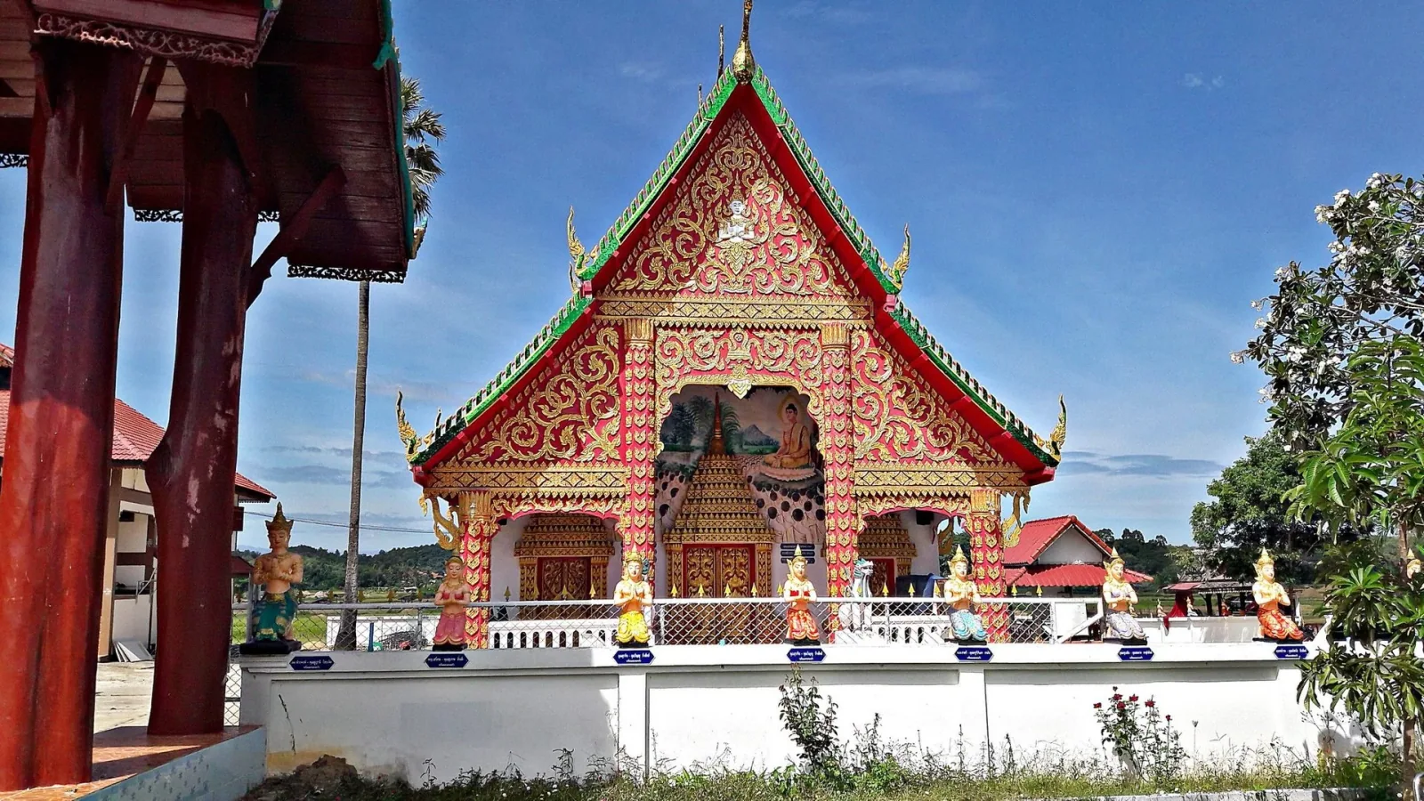 A vibrant Thai temple with a red and gold ornate facade, green roof trim, and multiple statues along a white wall in front, under a clear blue sky.