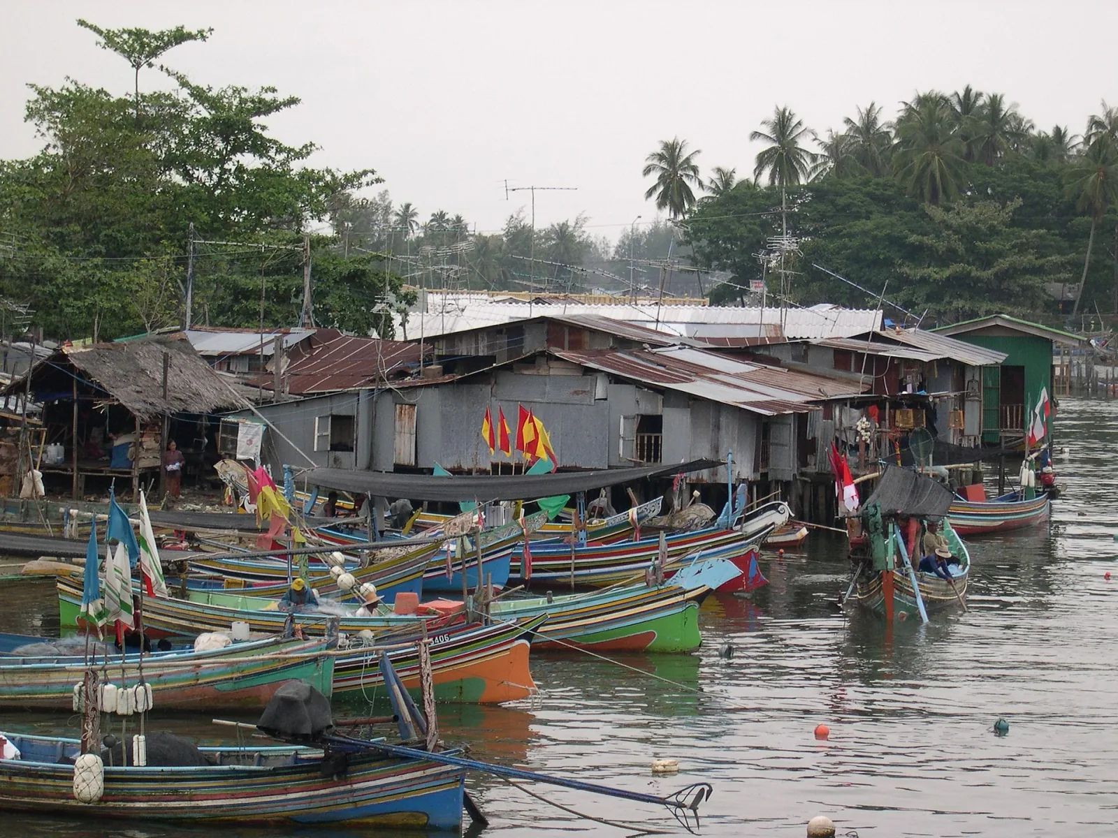 Colorful longtail fishing boats are moored in front of stilt houses in a bustling coastal village, with palm trees in the background.