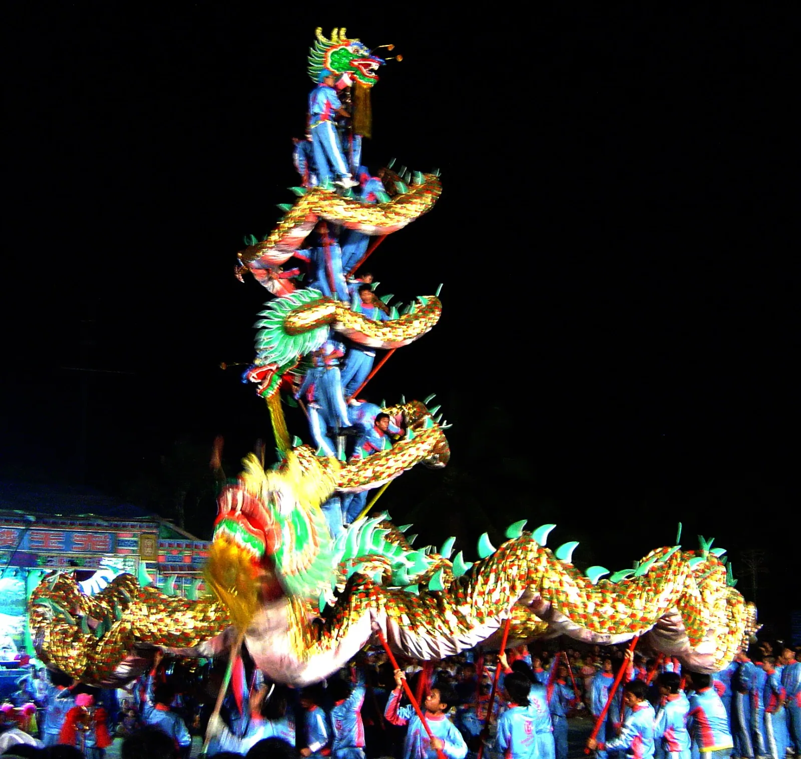 A long, golden Chinese dragon costume is held aloft by many performers at night, with several performers forming a human tower to elevate the dragon's head and upper body.