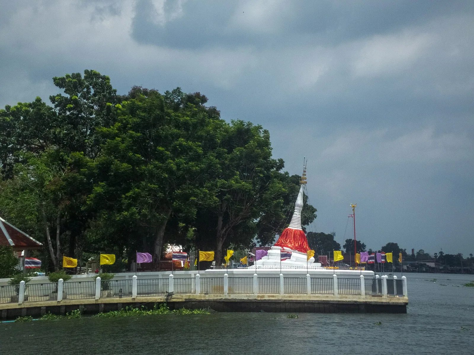 The leaning white Mutao Pagoda, adorned with a red sash, stands on a white-railed platform at the edge of a river, surrounded by green trees and colorful flags under a cloudy sky.