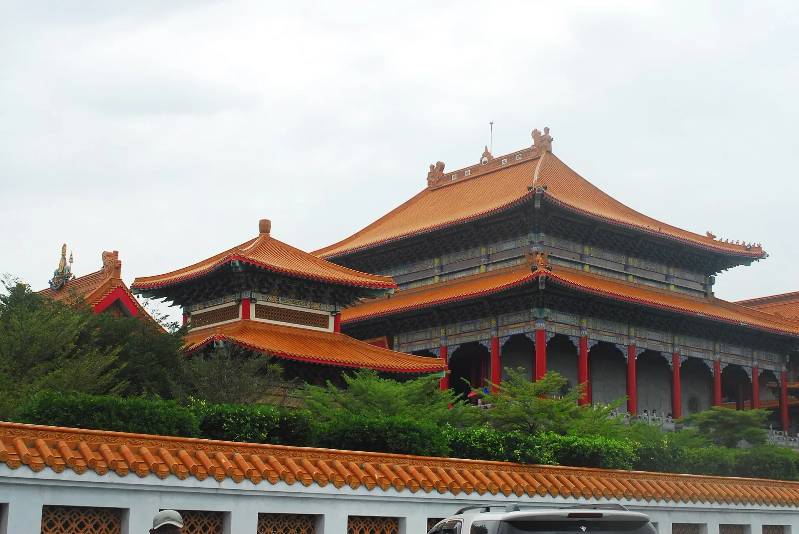 Wat Borom Racha Kanchana Phi Sek Anuson, a temple in Nonthaburi, featuring traditional Chinese-style architecture with orange tiled roofs and red pillars, partially obscured by trees and a decorative wall.