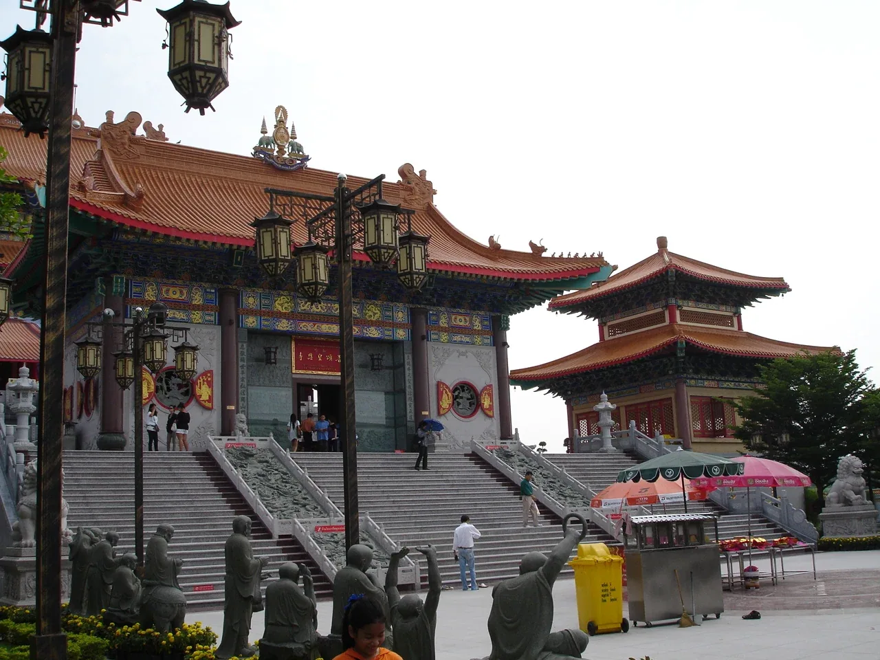 Exterior view of Wat Boromracha Kanchanapisek Anusorn, a grand Thai-Chinese temple with a multi-tiered orange roof, intricate facade, and a wide staircase leading to its entrance. Stone statues and decorative lanterns are visible in the foreground.