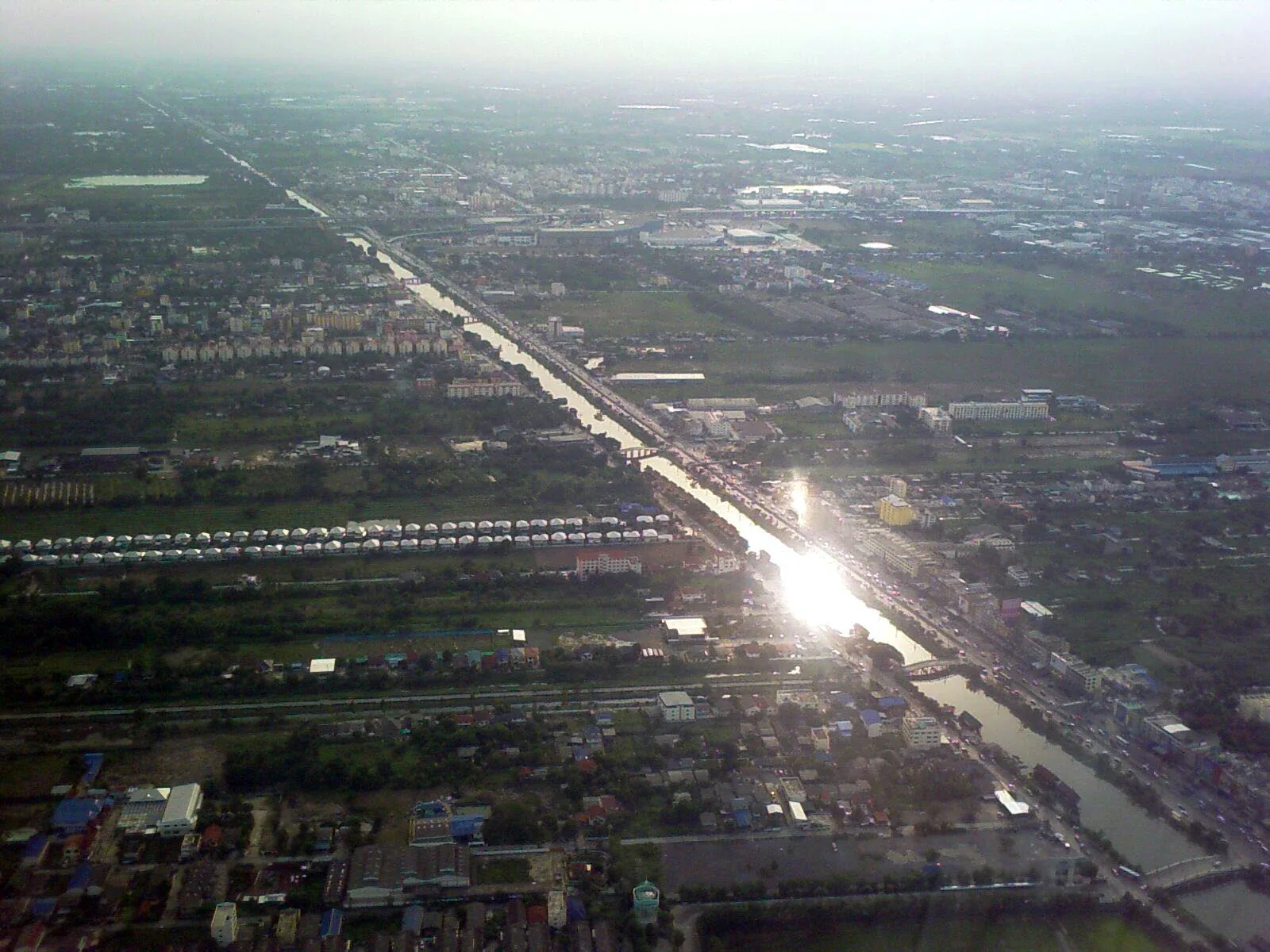 Aerial view of the Rangsit Canal in Pathum Thani, Thailand, with bright sunlight reflecting off its surface, surrounded by urban development and some green areas.