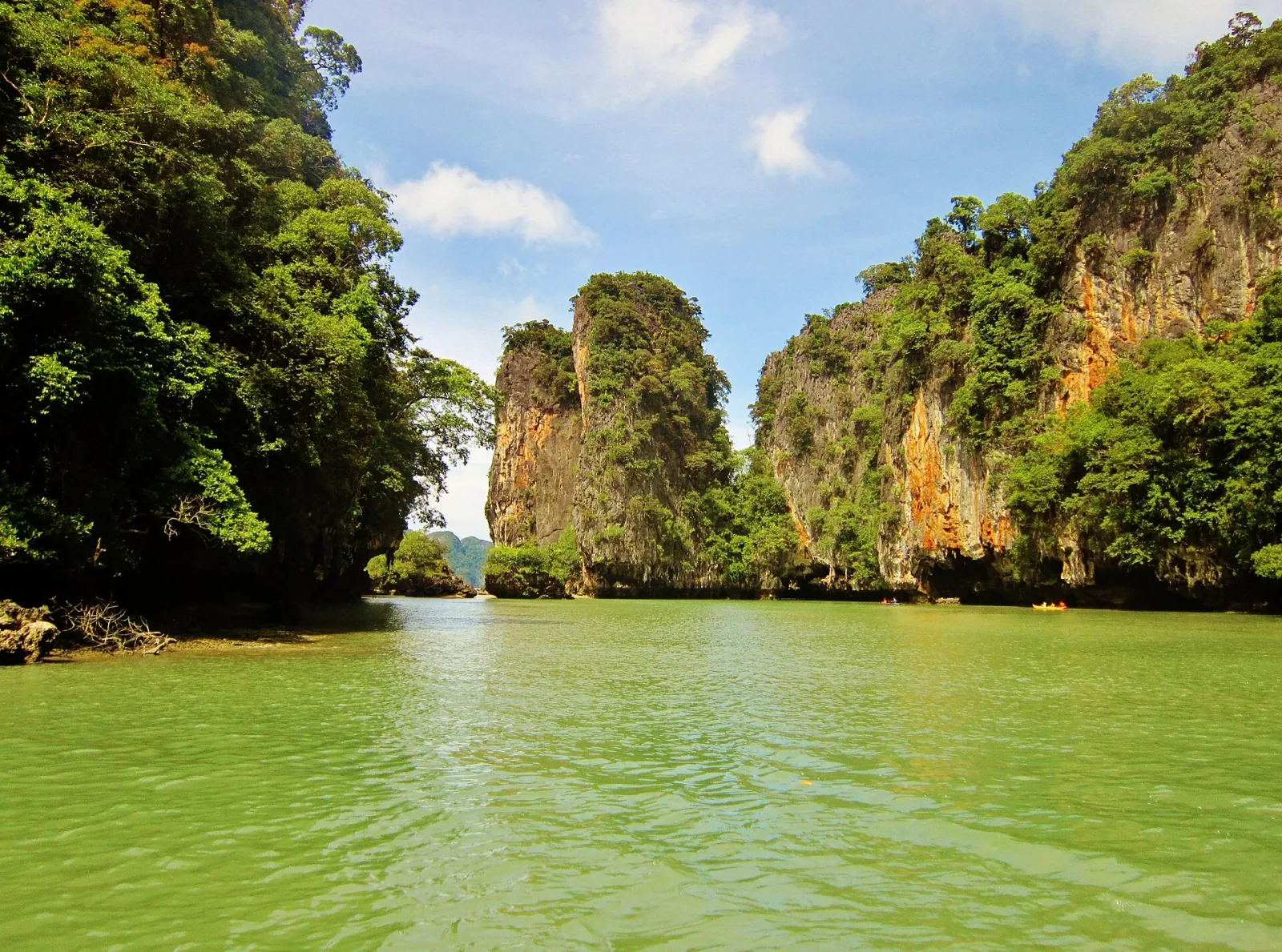 Towering, jungle-covered limestone karsts rise dramatically from the calm, green waters of Phang Nga Bay under a bright blue sky with scattered clouds.