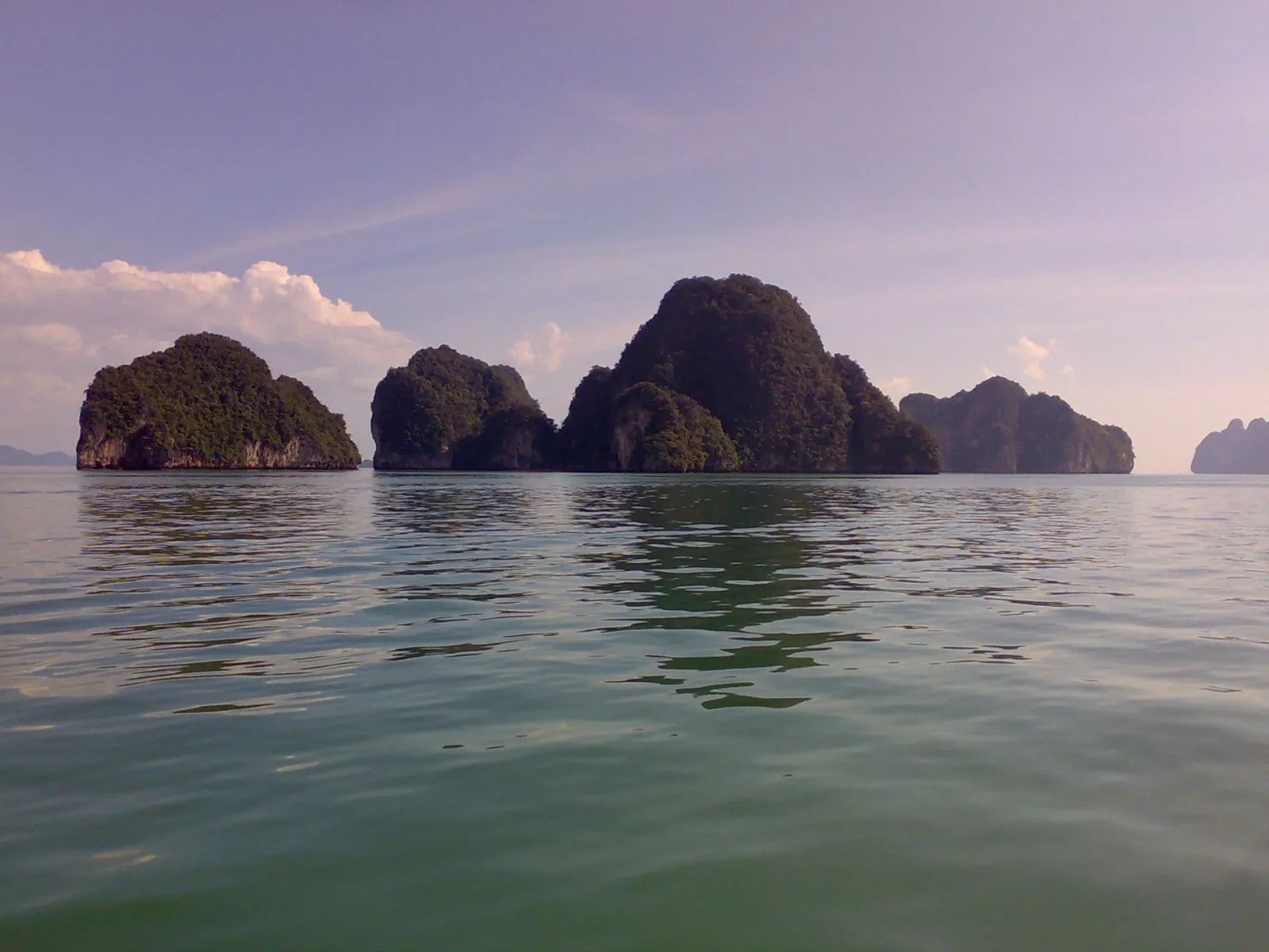 View of several large, tree-covered limestone islands emerging from the calm, green-blue waters of Phang Nga Bay under a light blue sky with scattered white clouds.