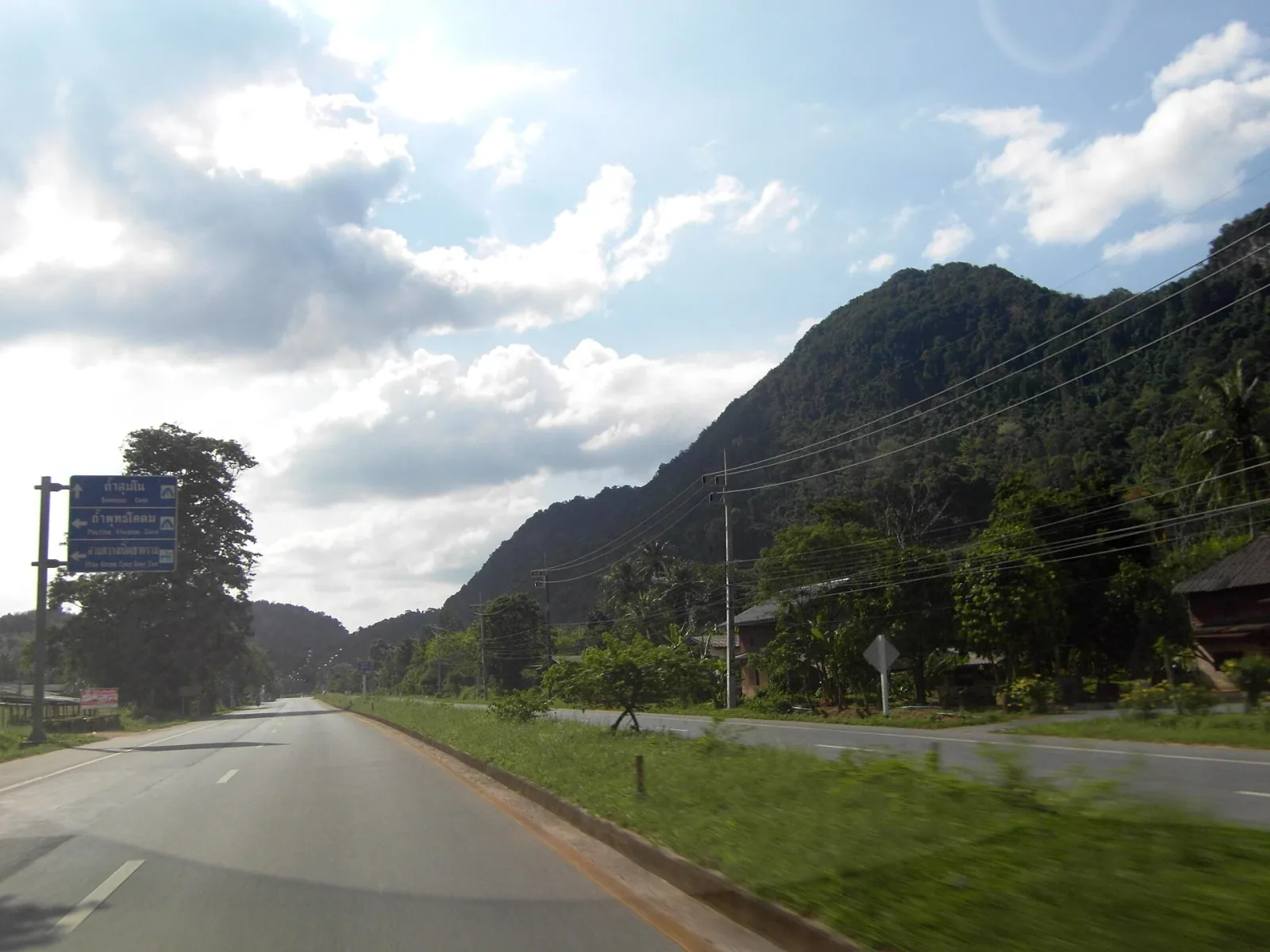 A two-lane road winding through a mountainous, tree-covered landscape in Phatthalung, Thailand, with a blue road sign visible on the left.