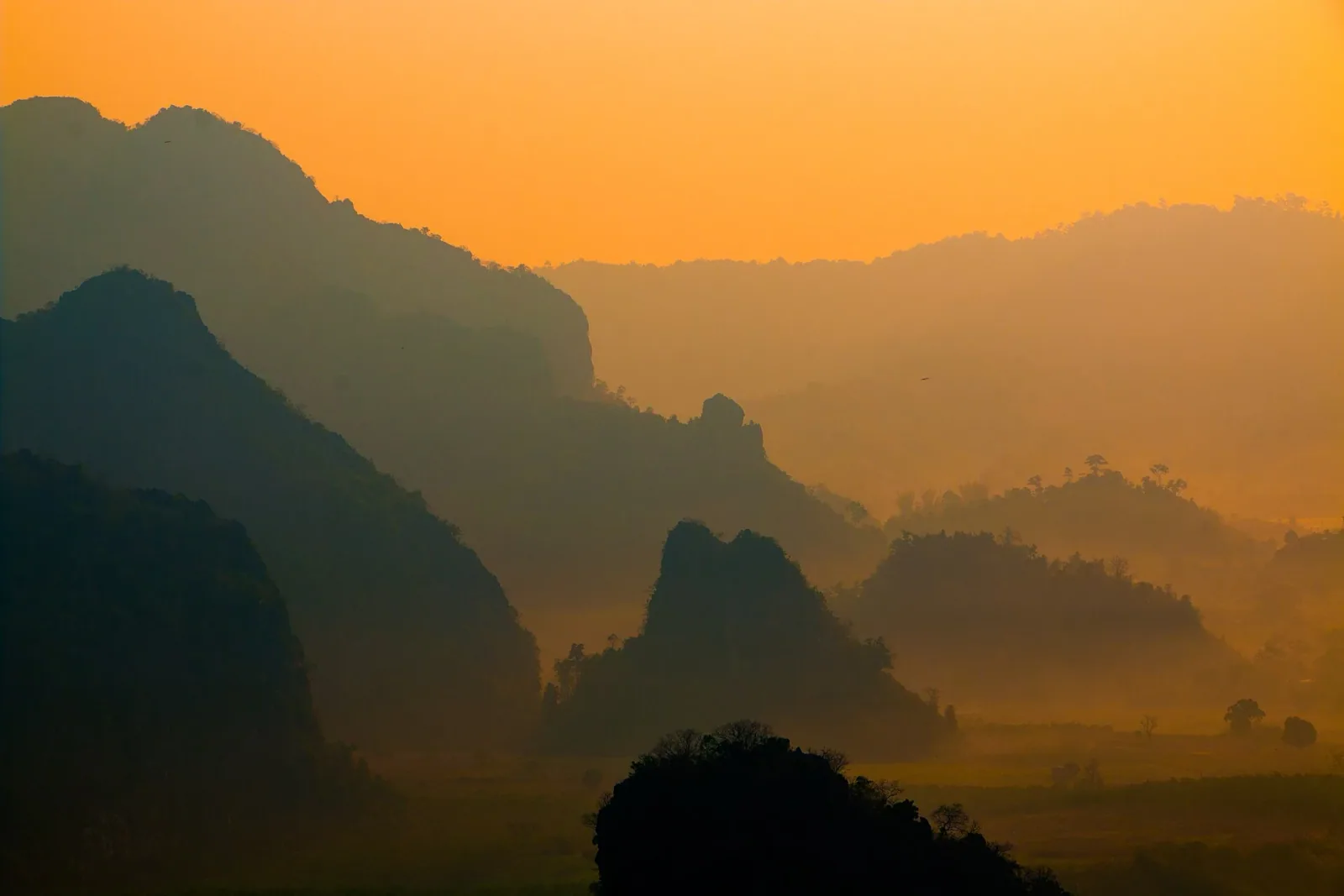 Layered mountain ranges silhouetted against a vibrant orange and yellow sky, with atmospheric haze creating depth.