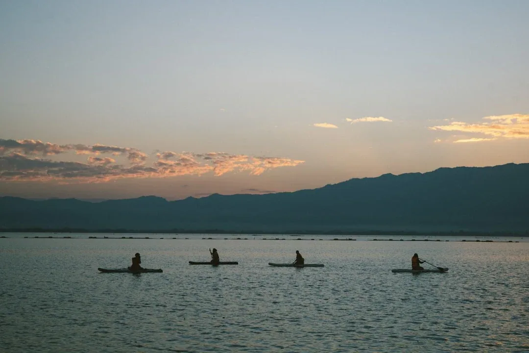 Four people in kayaks or canoes on Kwan Phayao lake at sunset, silhouetted against the water and distant mountains.