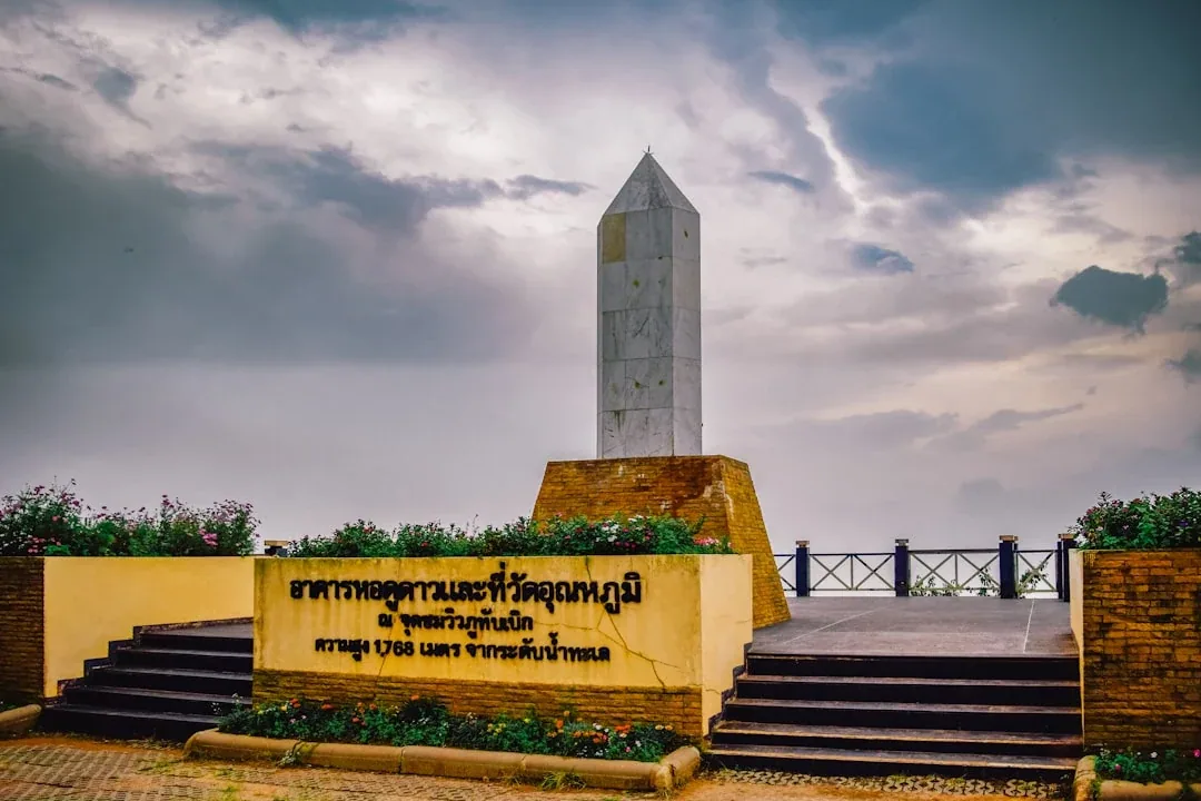 The monument at Phu Thap Boek viewpoint in Phetchabun, Thailand, with a sign indicating its elevation and purpose, under a cloudy sky.