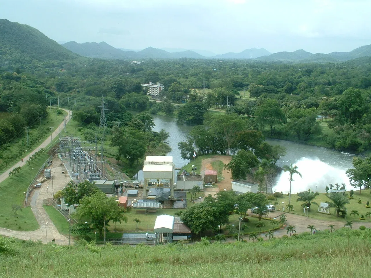 An aerial view of Kaeng Krachan Dam's hydroelectric power plant, with water gushing into the river, surrounded by lush green hills and forests under a cloudy sky.
